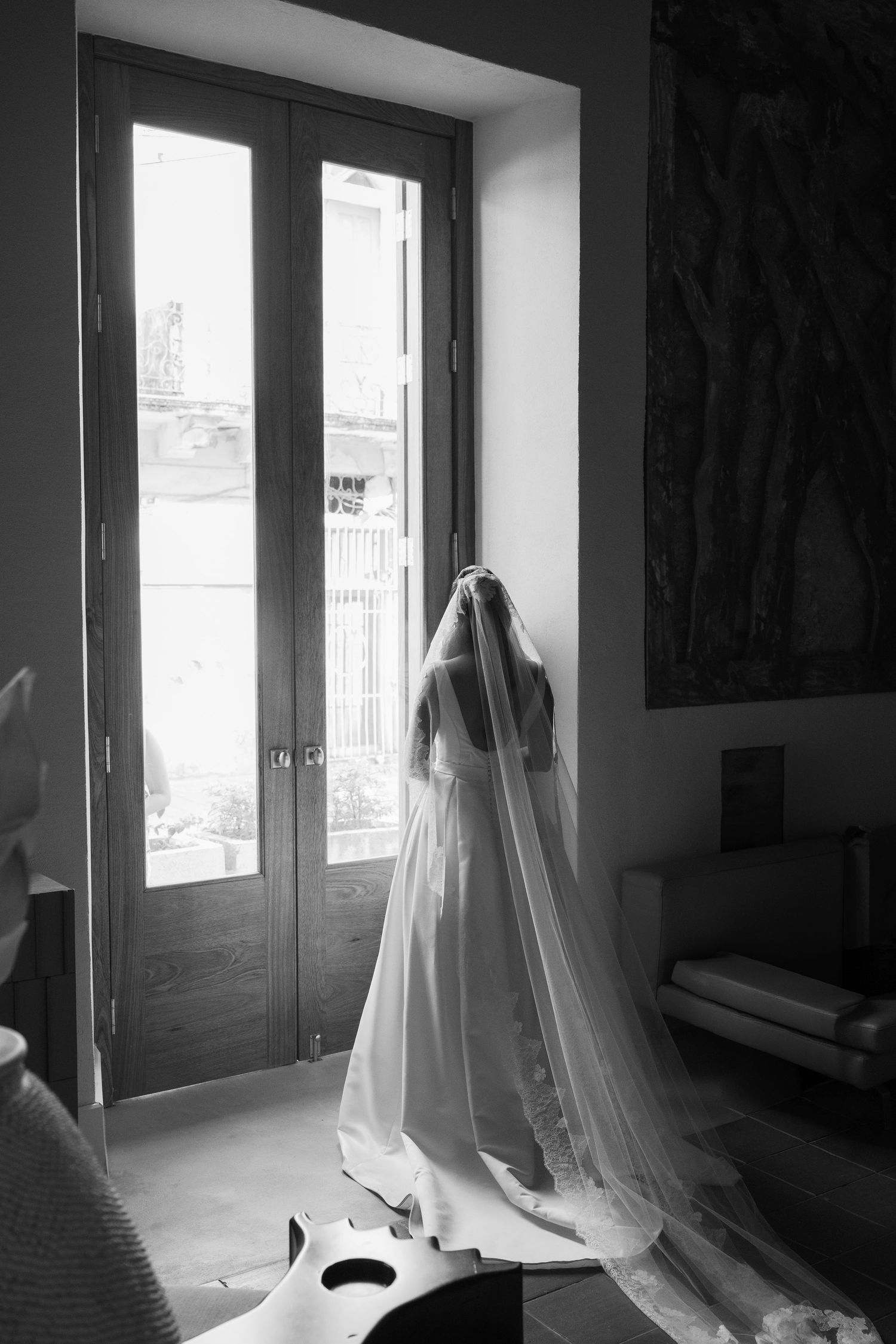 A black and white photograph of a bride in a flowing veil and dress standing by a tall window in natural light.