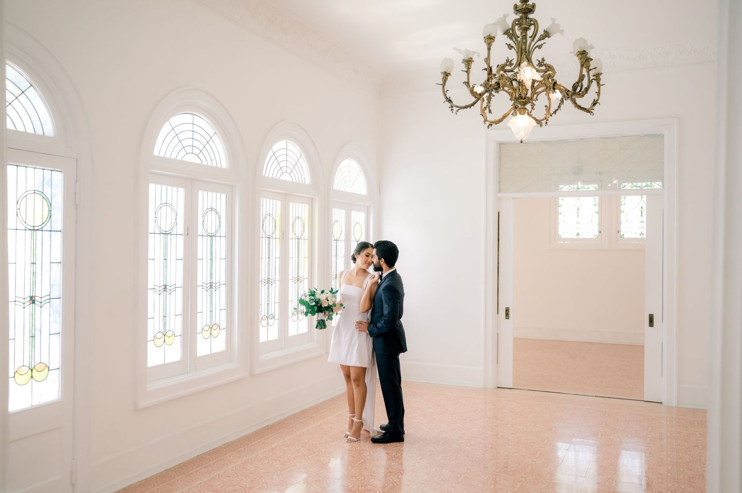 Bright white wedding venue interior with arched windows, chandelier and elegant architectural details.