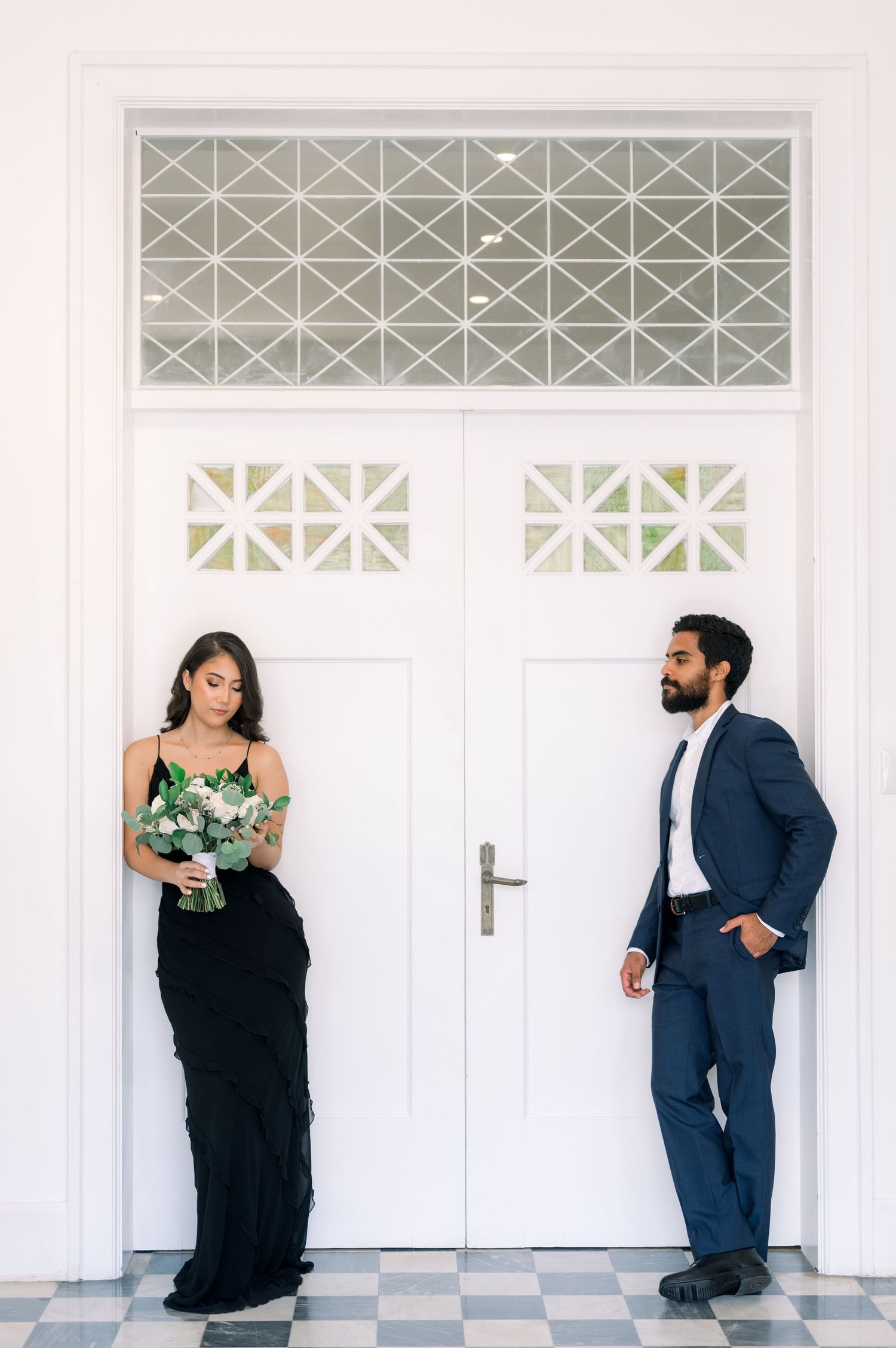 Elegant couple poses against white doors with geometric patterns, bride holding bouquet and groom in navy suit.