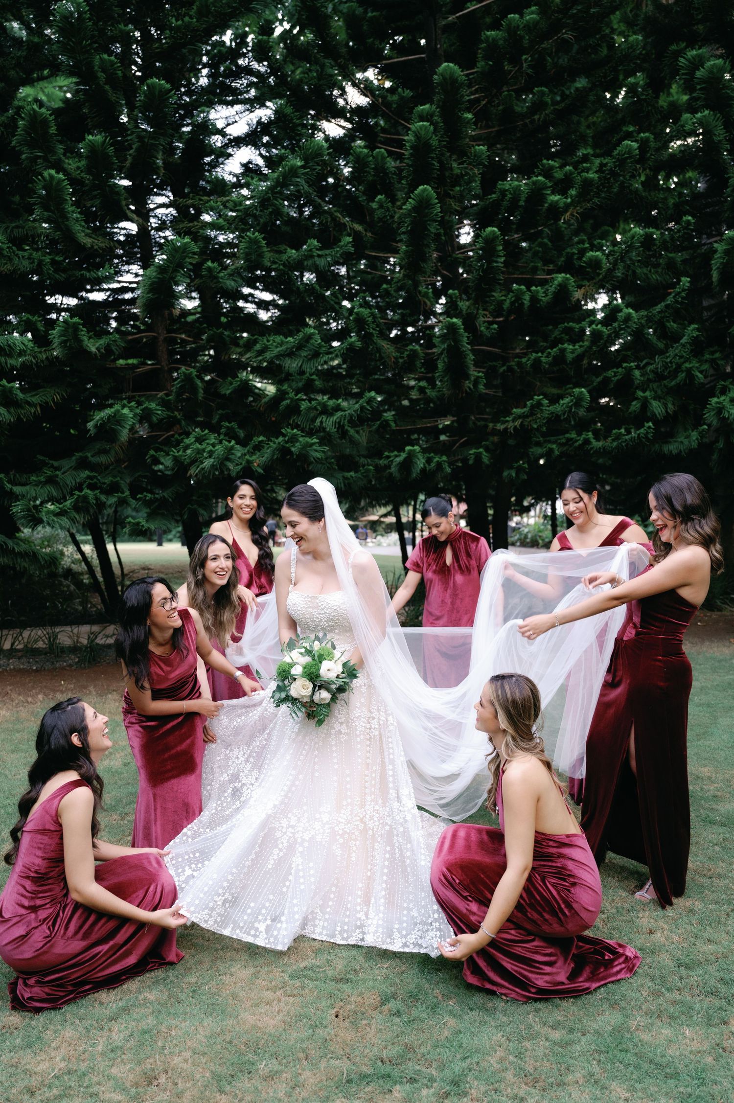 A wedding party in burgundy dresses helps arrange the bride's white lace dress in a flowing sequence of outdoor photos.