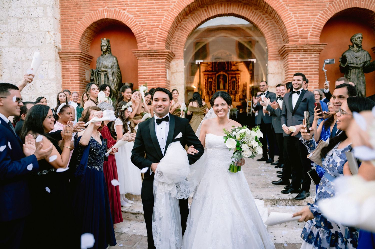 Newlyweds exit church ceremony while guests throw confetti and celebrate under brick archways.