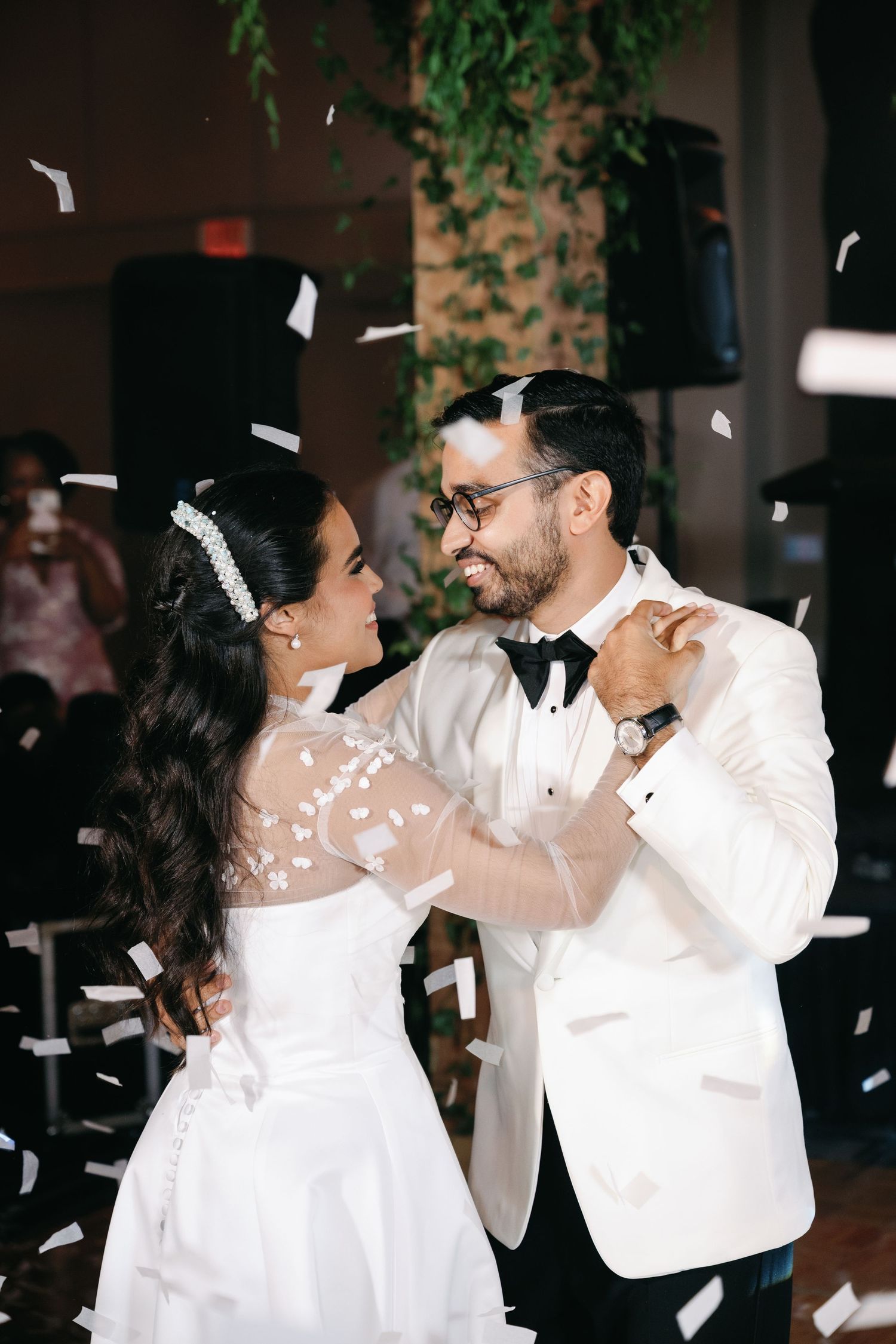 A couple in white formal attire dances together at a wedding reception as confetti falls around them.