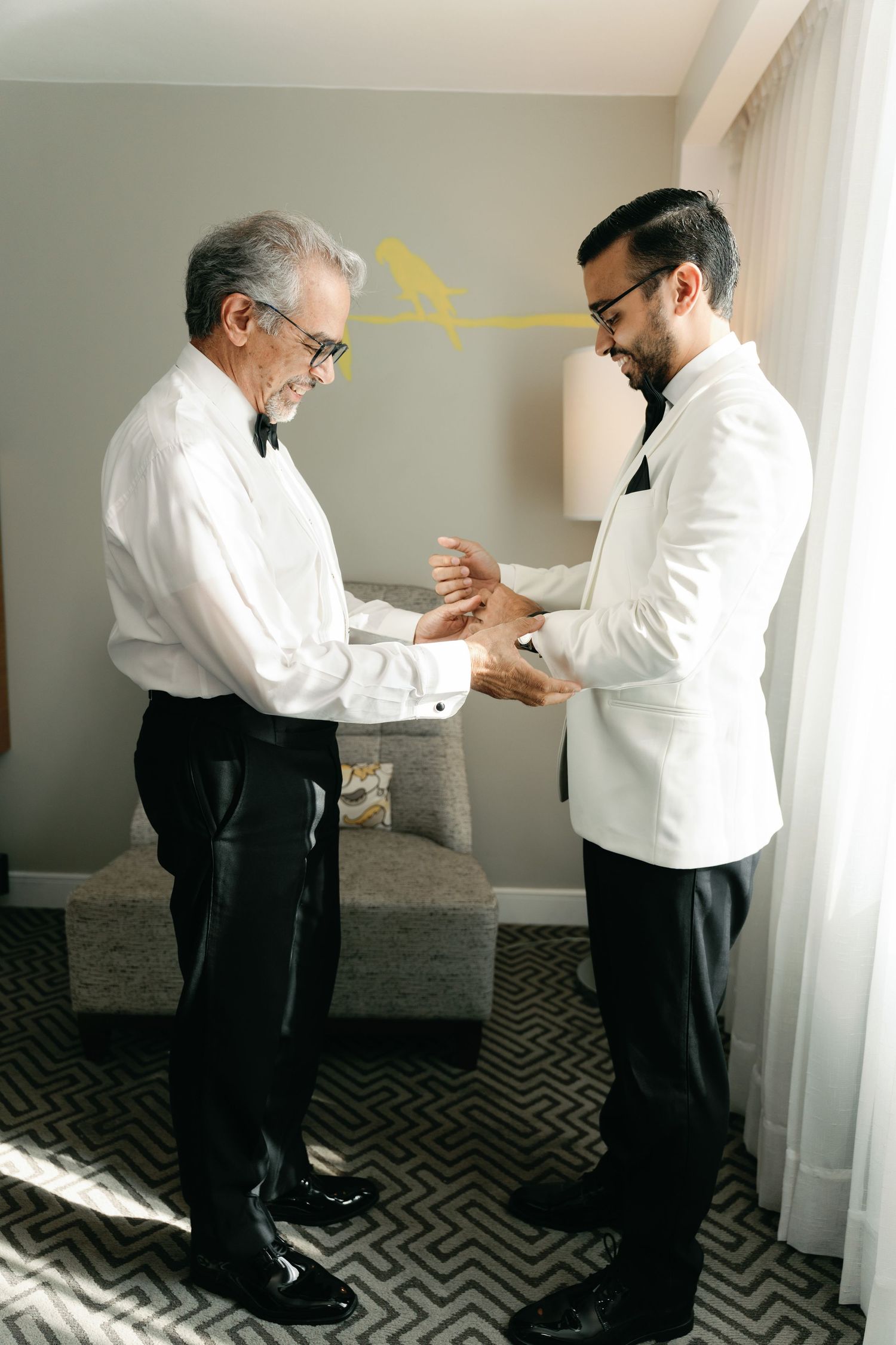 Two men in formal attire help each other with cufflinks and final adjustments while getting ready in a hotel room.