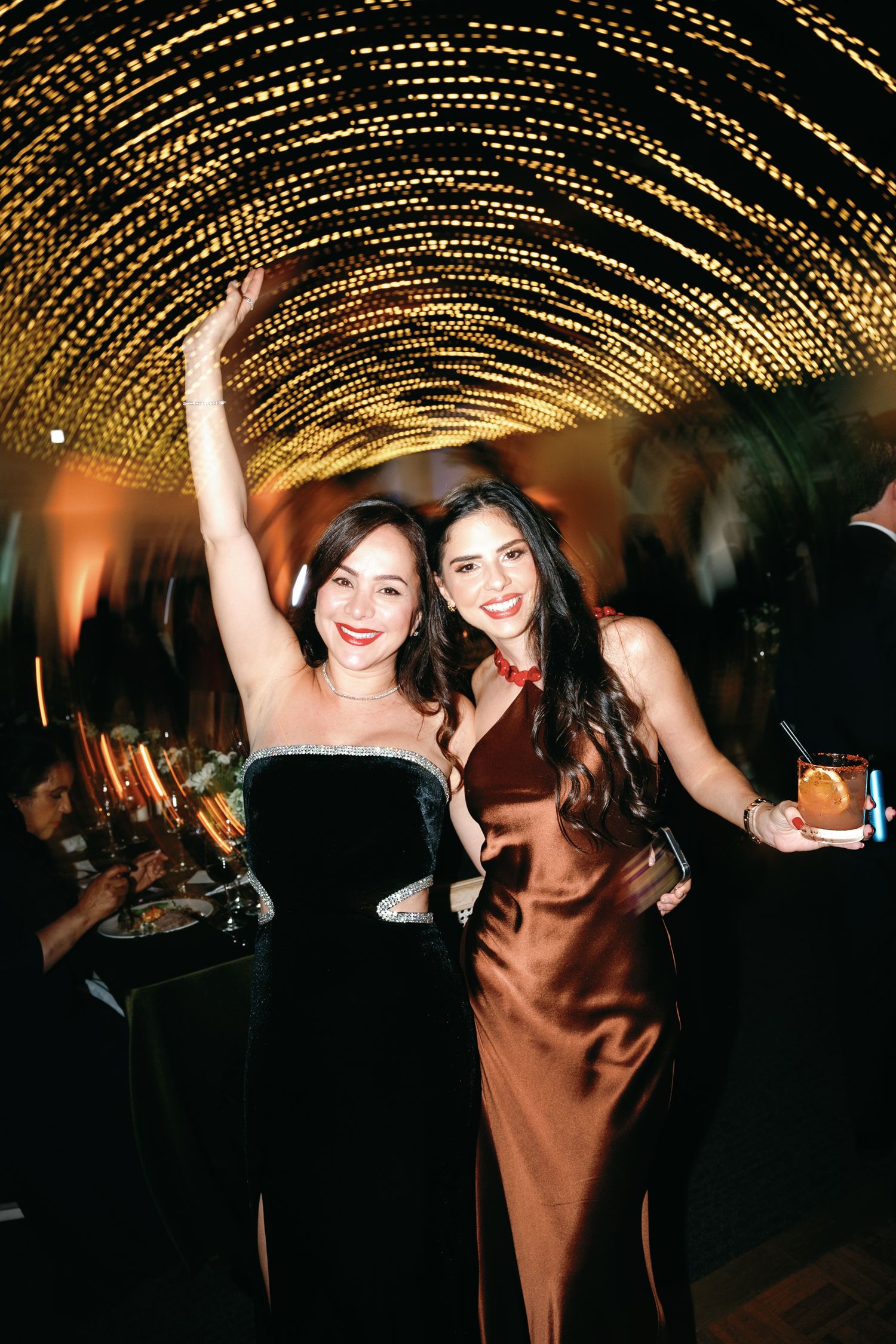 Two friends celebrate at an elegant party under glittering string lights in formal evening wear.