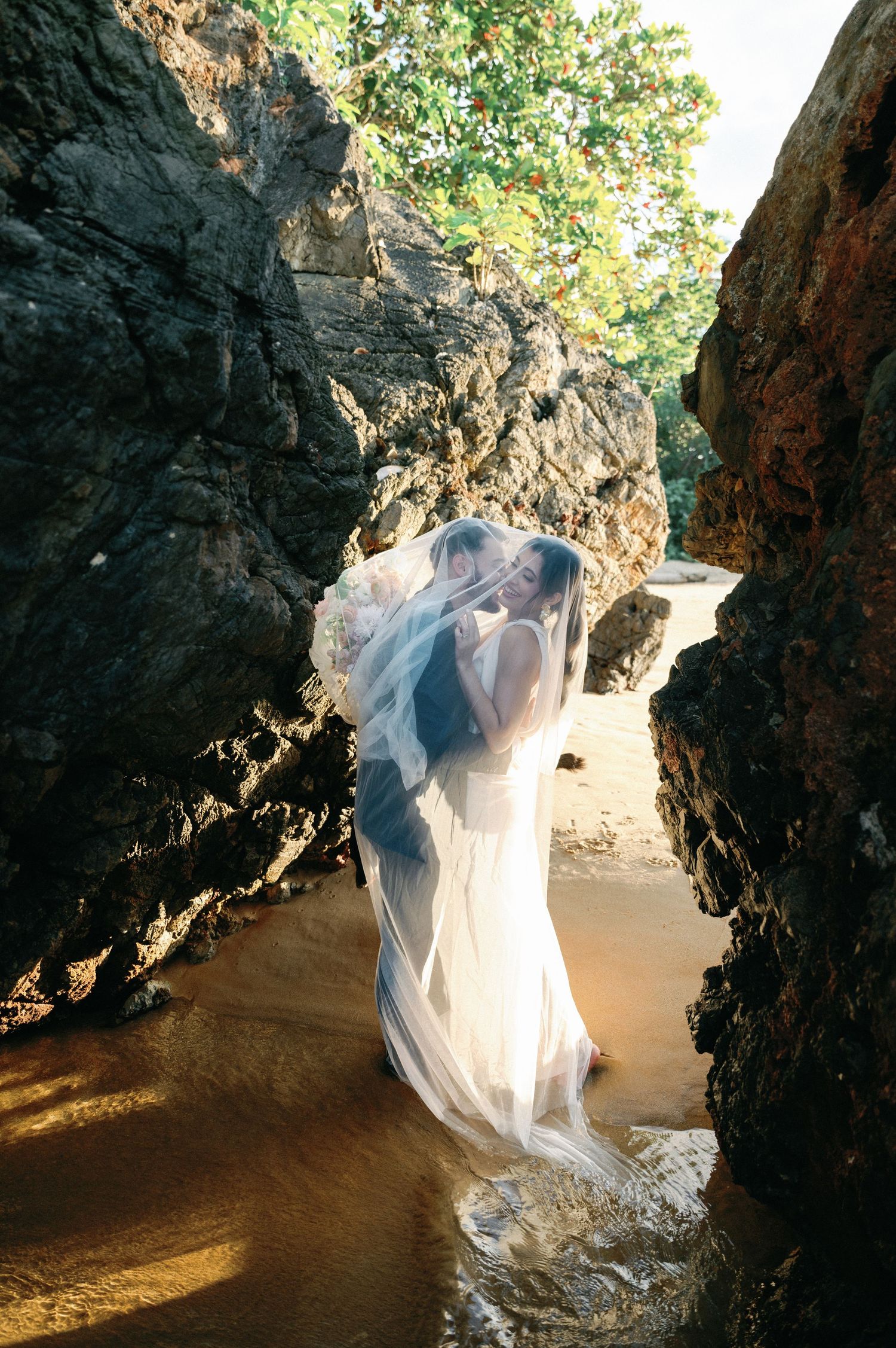 A flowing white veil catches sunlight between rocky cliffs on a beach, creating an ethereal scene.