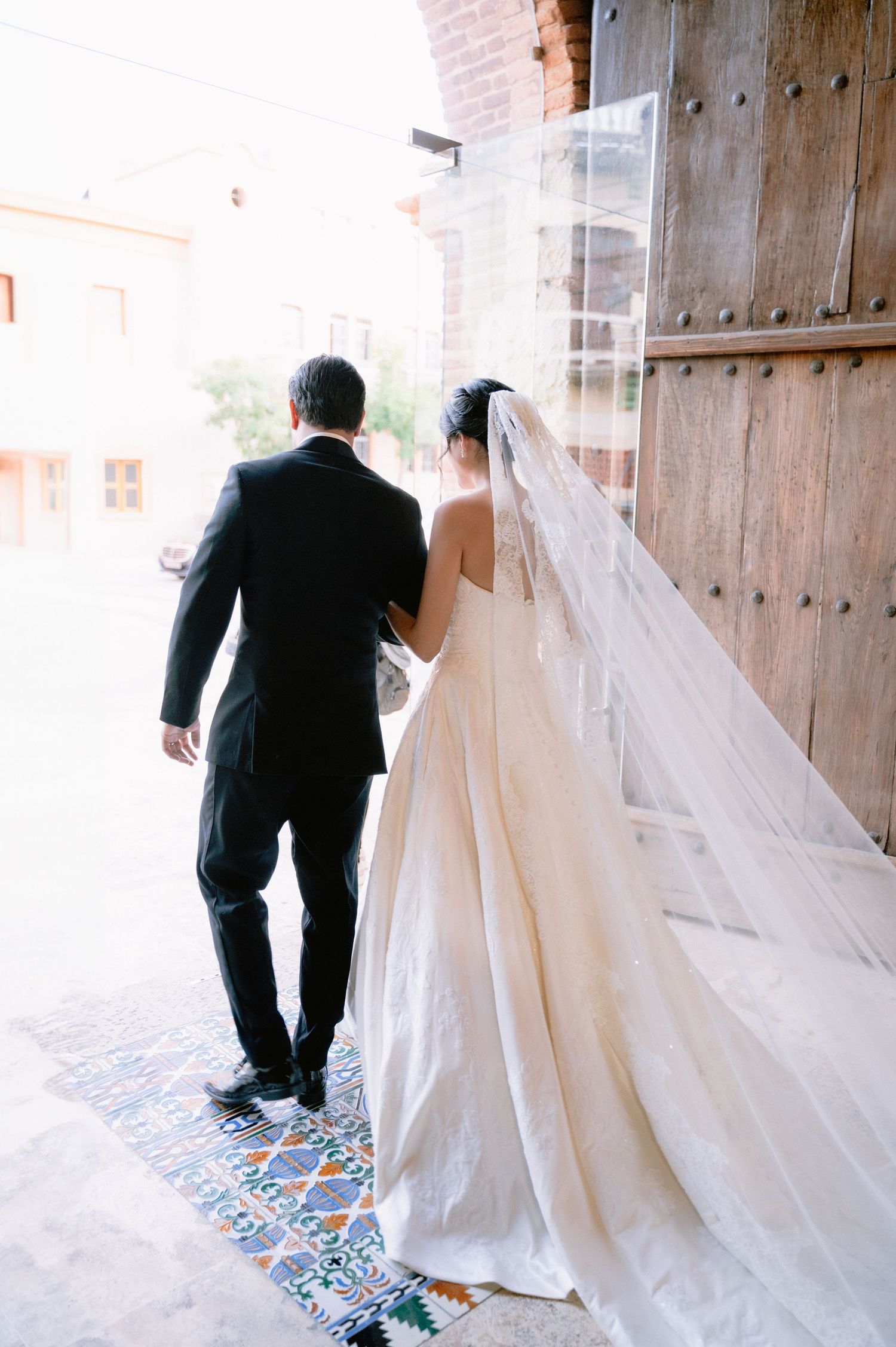 Long flowing wedding veil trails behind couple walking through rustic wooden doorway.