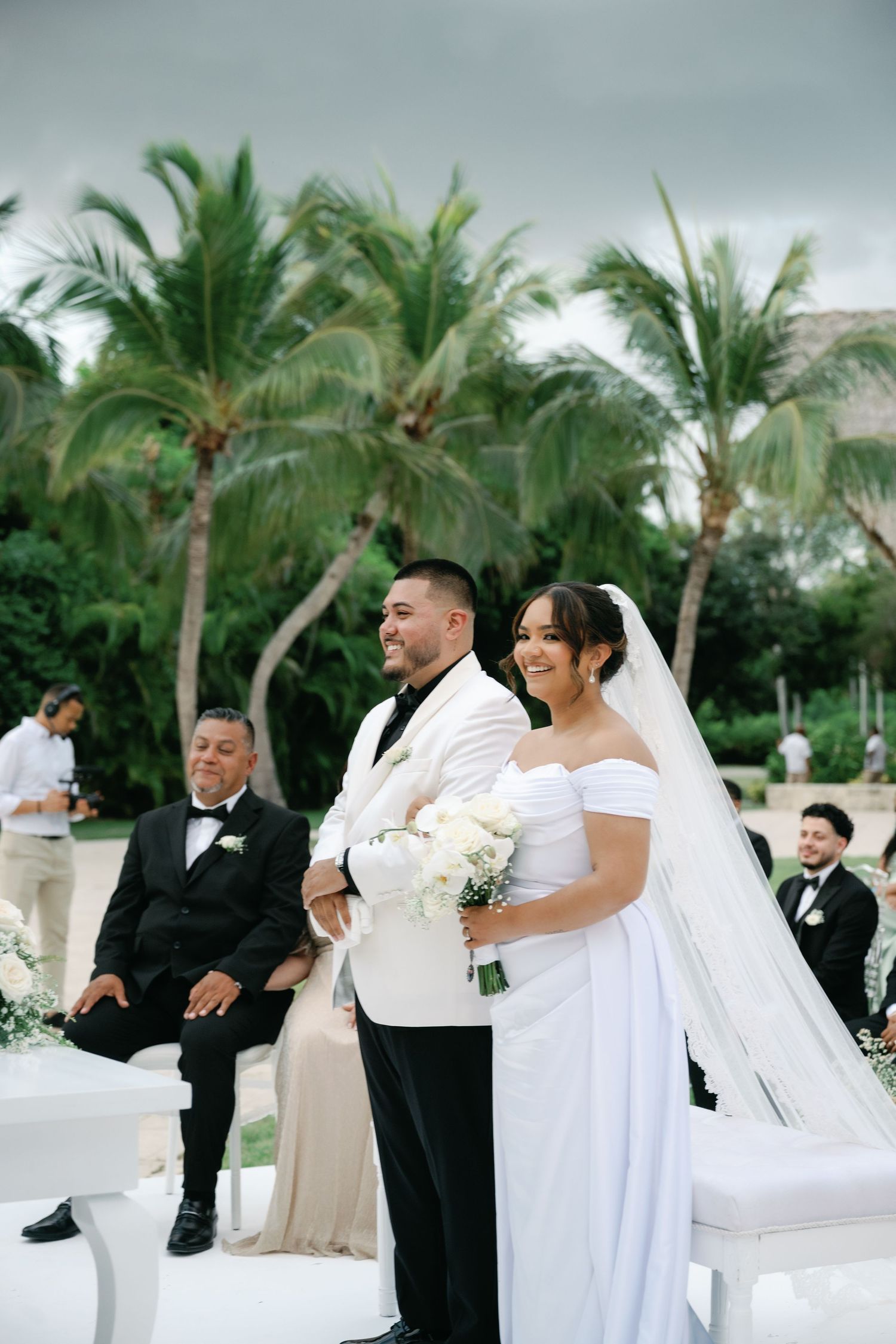 A wedding party celebrates outdoors with palm trees in the background during a tropical destination ceremony.