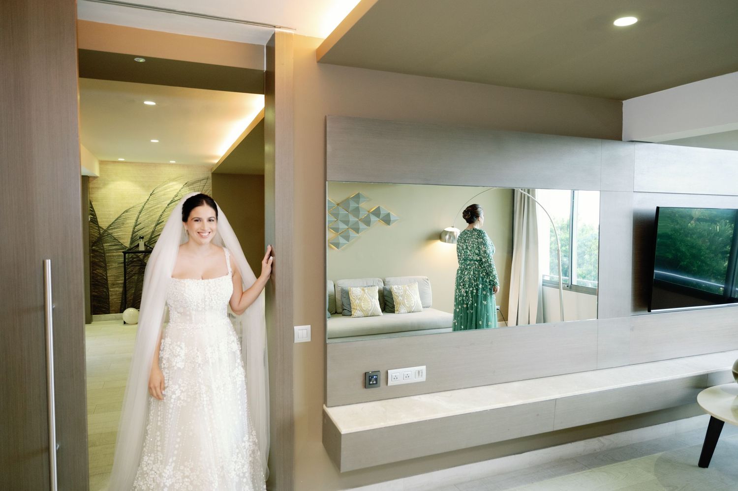 A bride in a white lace gown stands in a modern hallway with mirrors and sleek lighting.