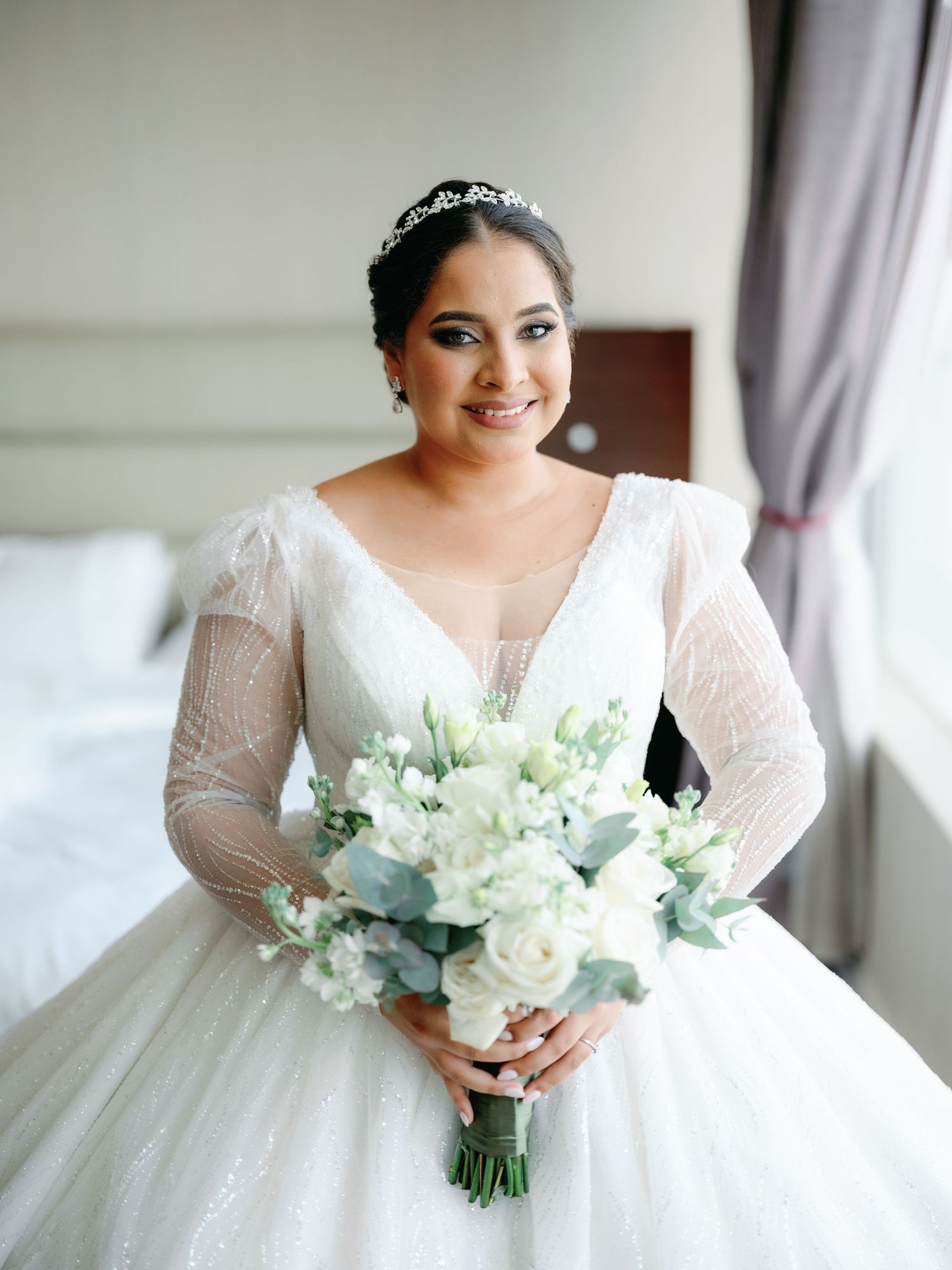 A bride in a white ball gown holds a delicate bouquet of white and green flowers against a light interior background.