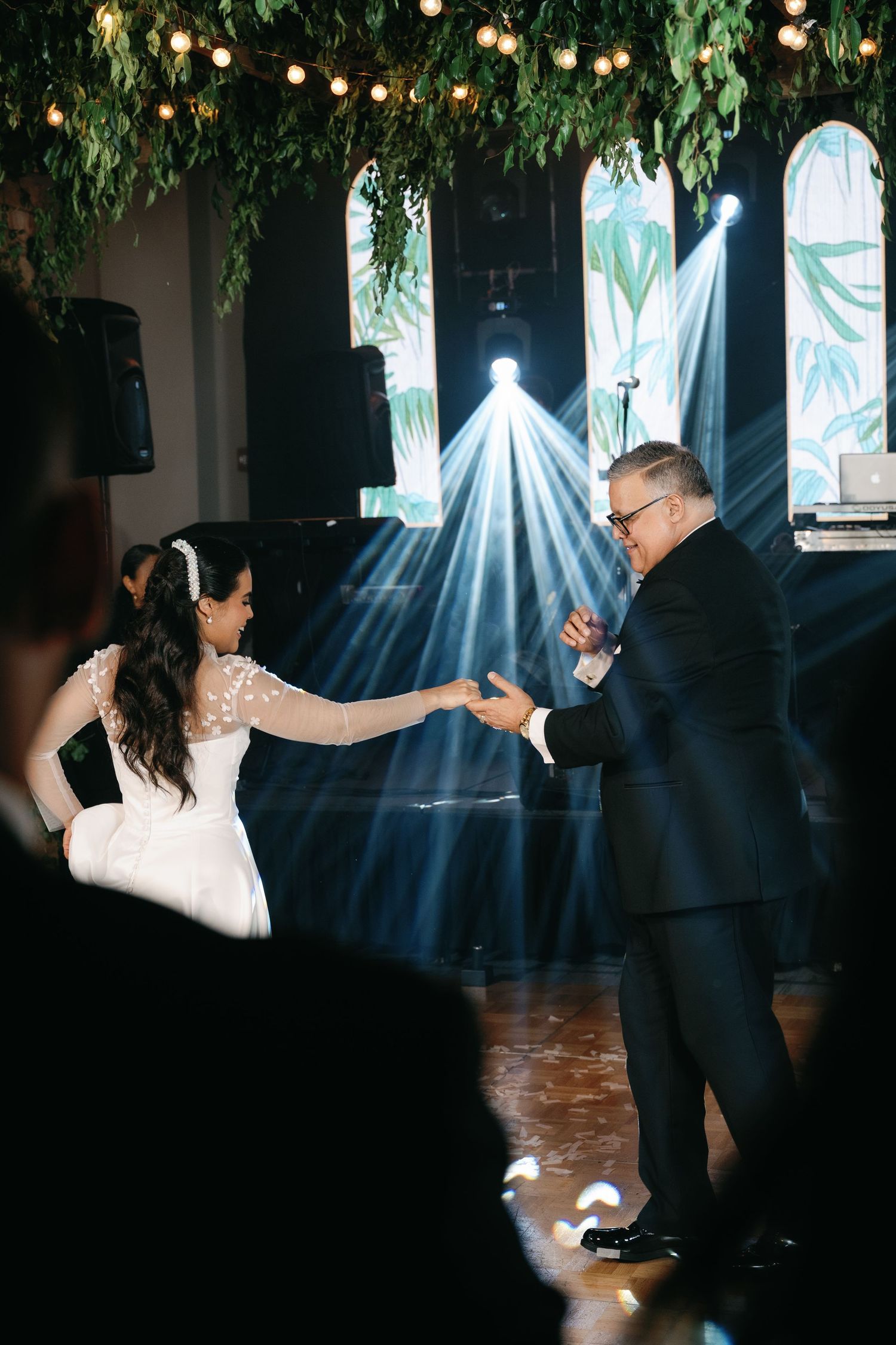A couple dances together in a dimly lit venue with string lights and greenery hanging overhead.