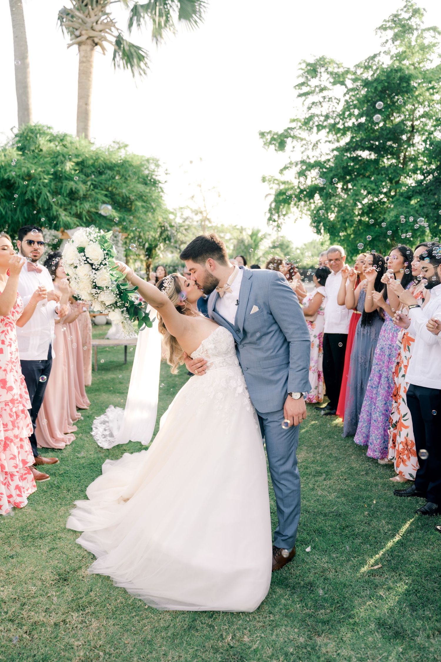A romantic wedding kiss on a lush green lawn with guests in colorful dresses and a palm tree backdrop.