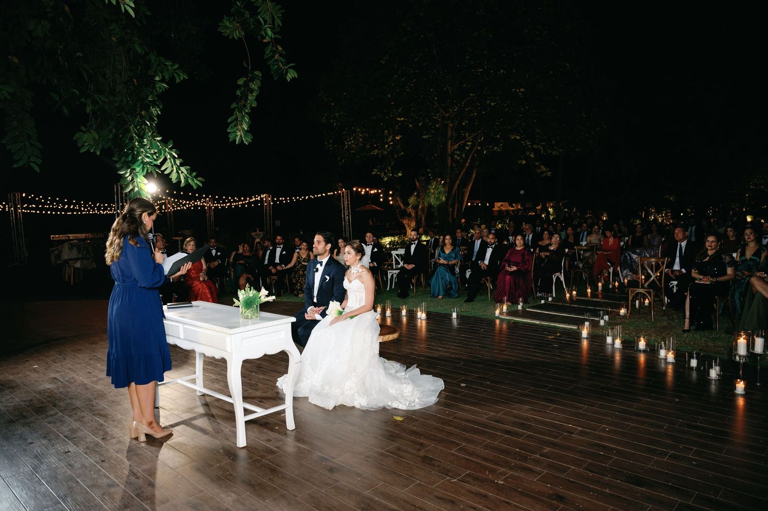 Wedding reception at night with string lights illuminating an outdoor venue and guests mingling in the background.