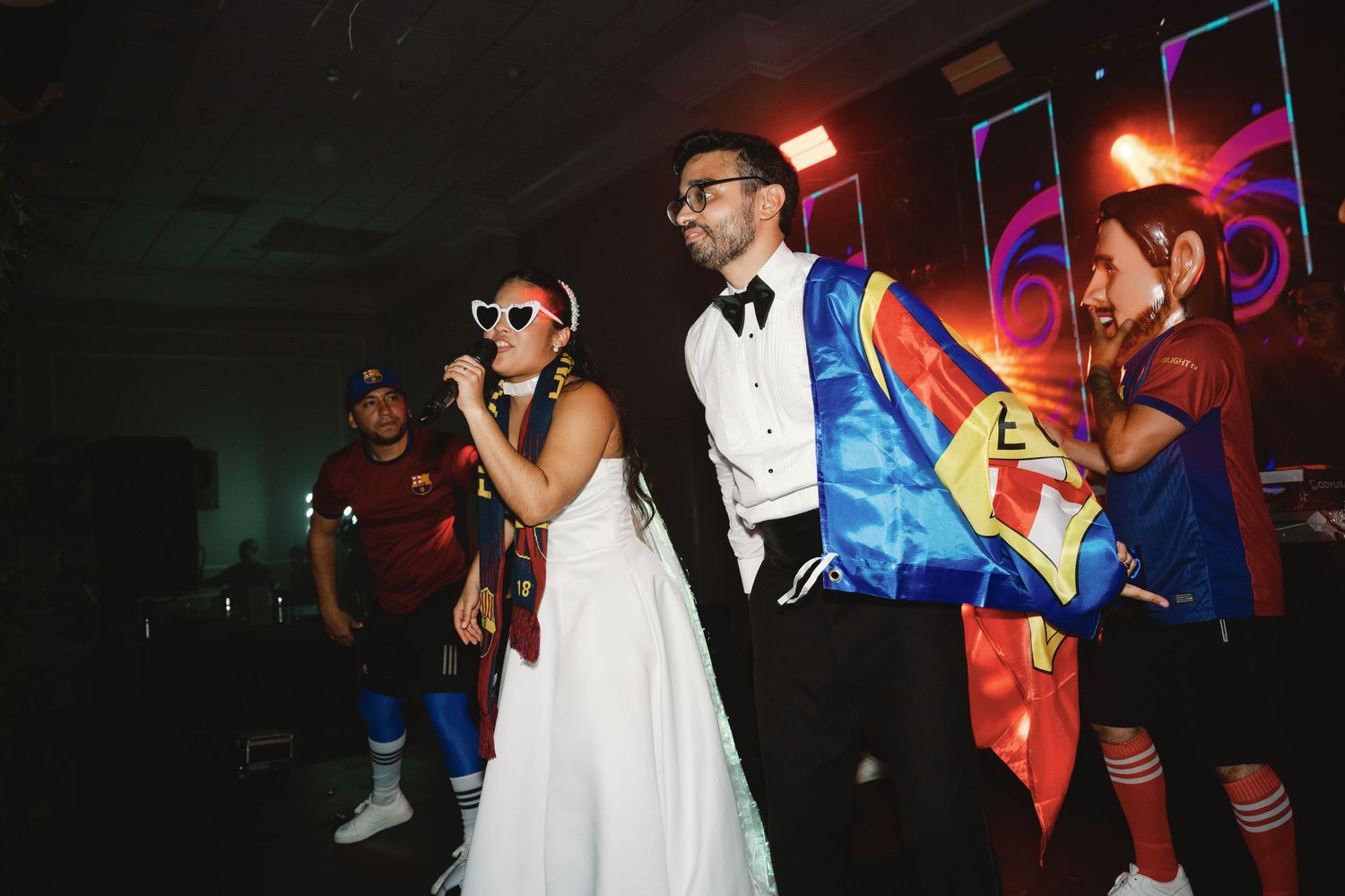 People dancing and singing at a wedding reception with colorful lighting and festive party attire.