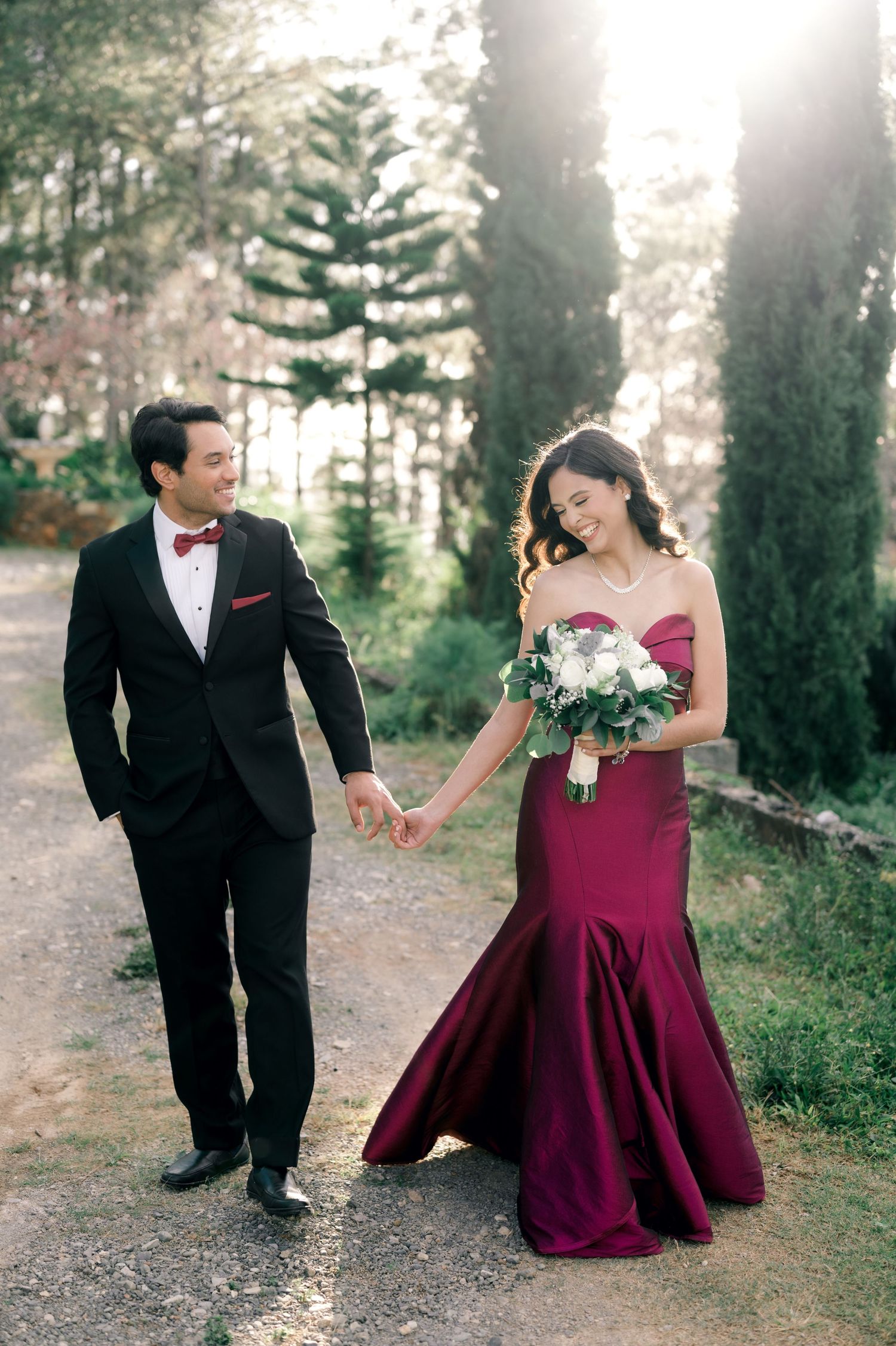 A couple in formal attire, with a burgundy gown and black tuxedo, share candid moments in a sunlit outdoor setting.