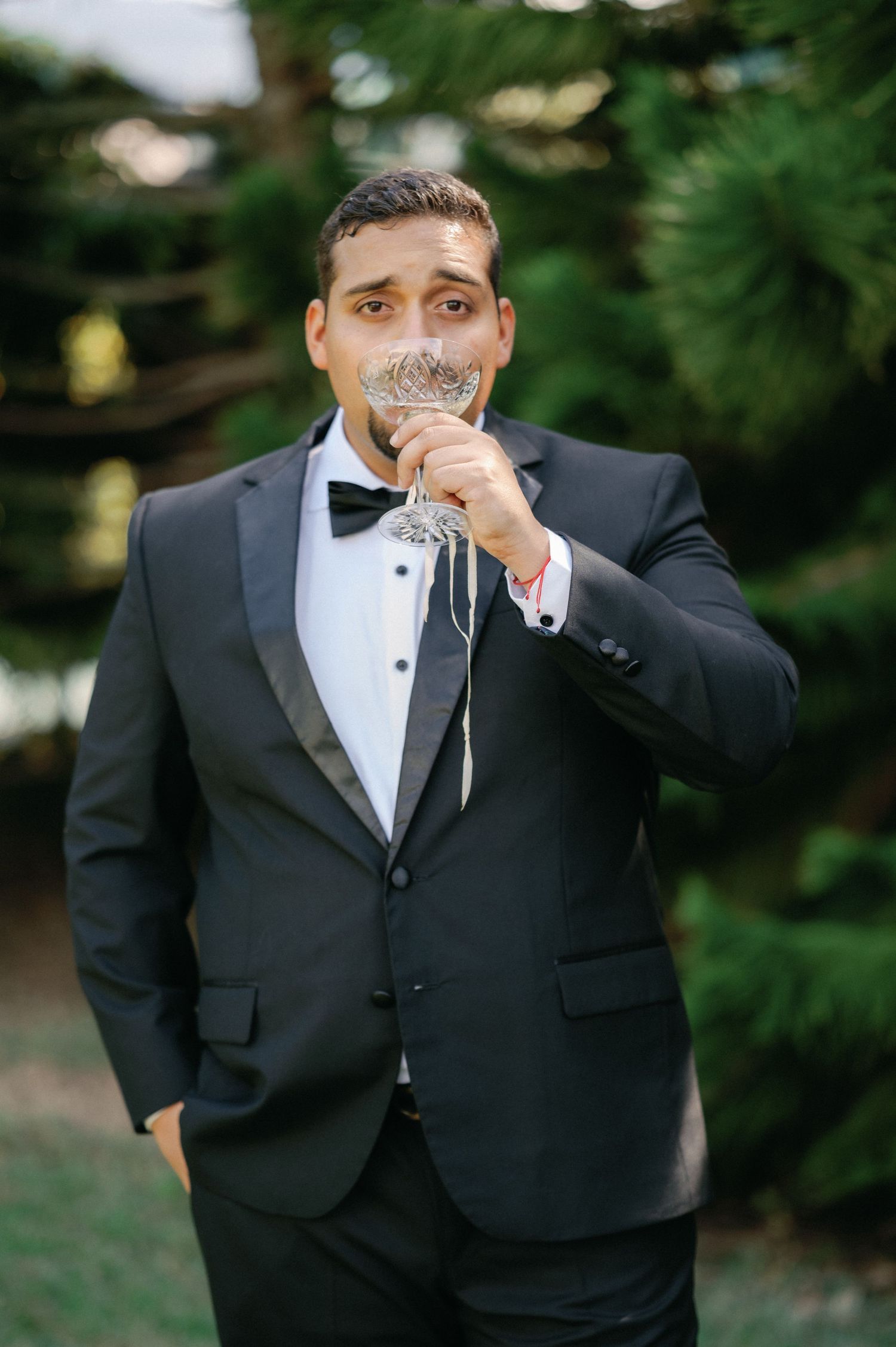 Person in formal black tuxedo and bowtie drinking from an elegant champagne glass outdoors.