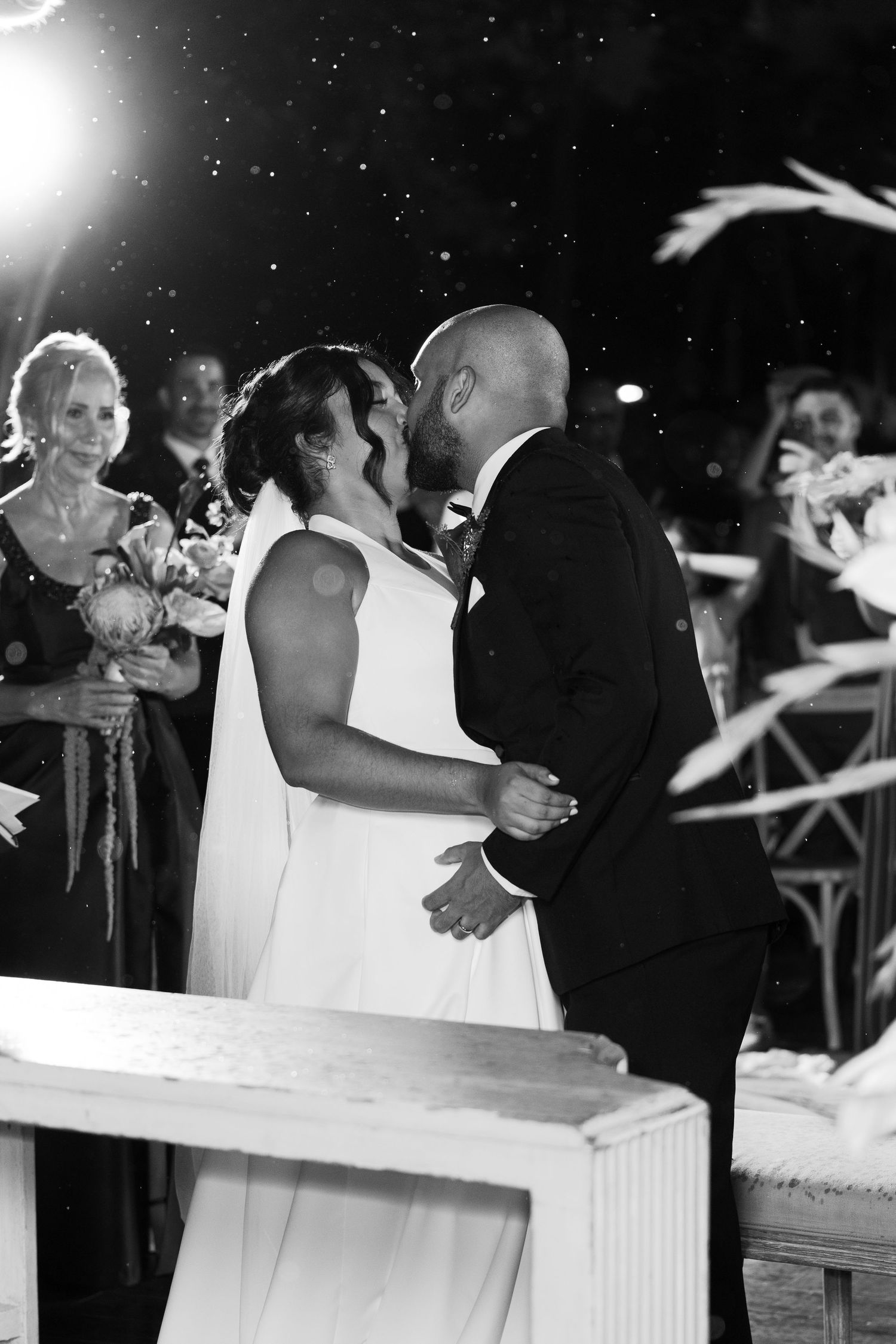 Wedding guests dance and celebrate at an outdoor evening reception with a black and white filter effect.