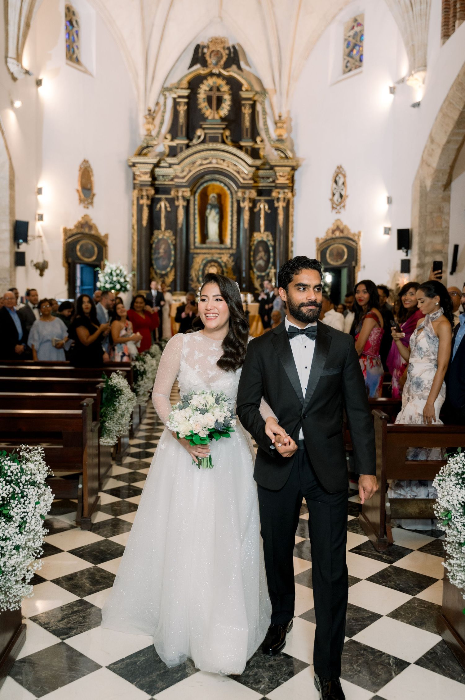 A wedding ceremony takes place in an elegant church with ornate decorations and a black and white checkered floor.