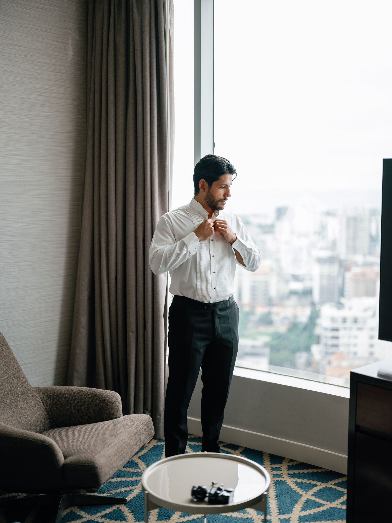 Professional adjusting shirt while standing by large window in high-rise hotel room with city view and modern decor.