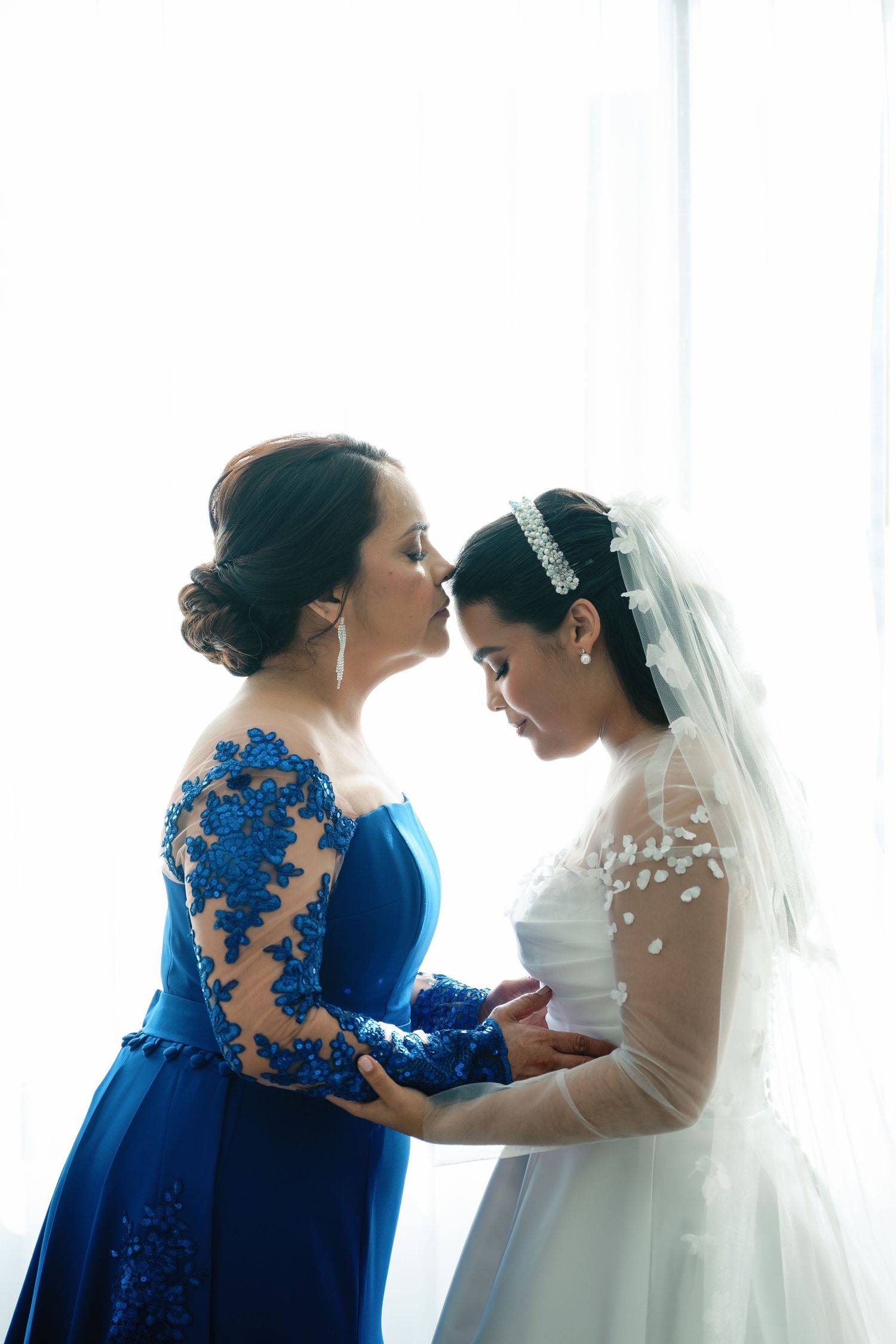 A tender moment between two women in formal dresses, one in royal blue lace and one in a white wedding gown with veil.