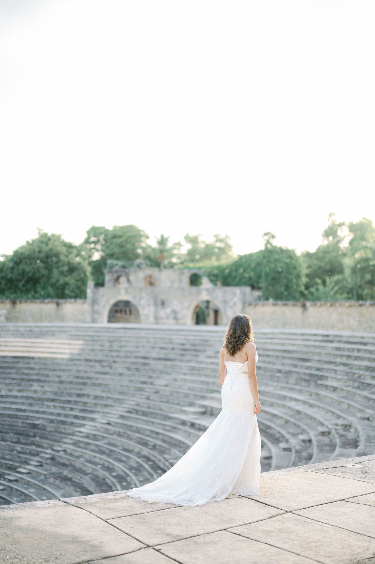 A bride in a flowing white wedding dress poses gracefully at an ancient stone amphitheater with curved stone seating.