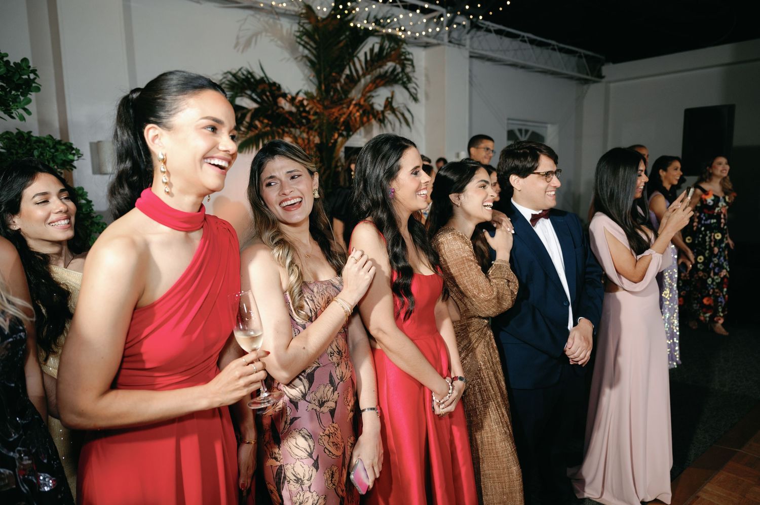 Bridesmaids in red and pink dresses stand together smiling and clapping during a wedding celebration.