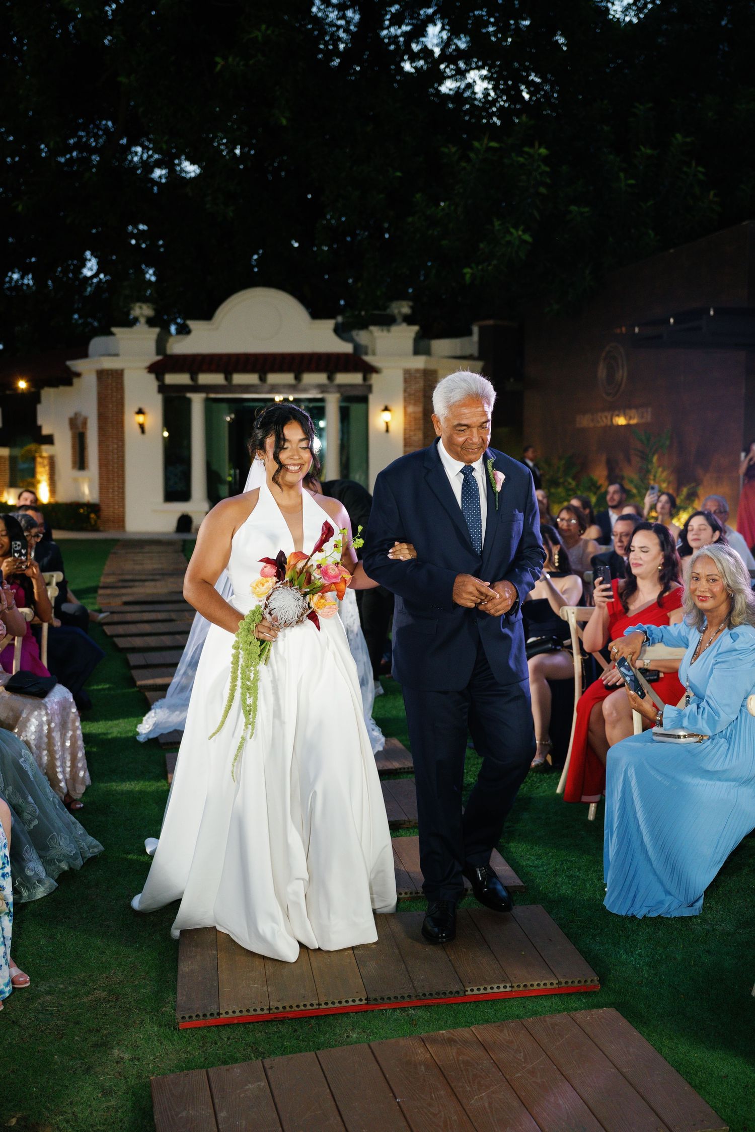 Wedding couple greets guests while walking down wooden deck pathway at outdoor evening reception.