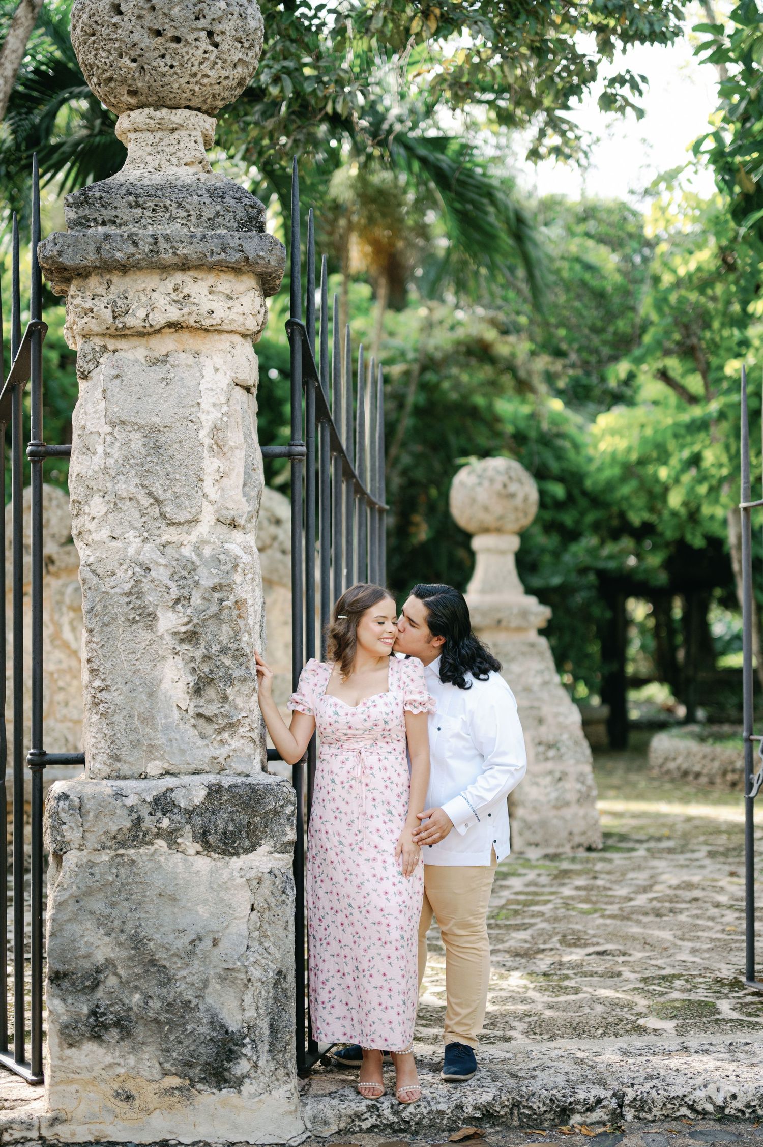 A romantic couple shares an intimate moment in front of a stone gate pillar in a garden setting.