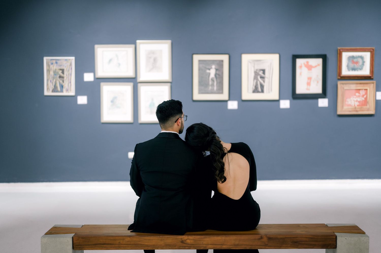 Two people sit together on a wooden bench viewing framed artwork displayed on a dark blue gallery wall.