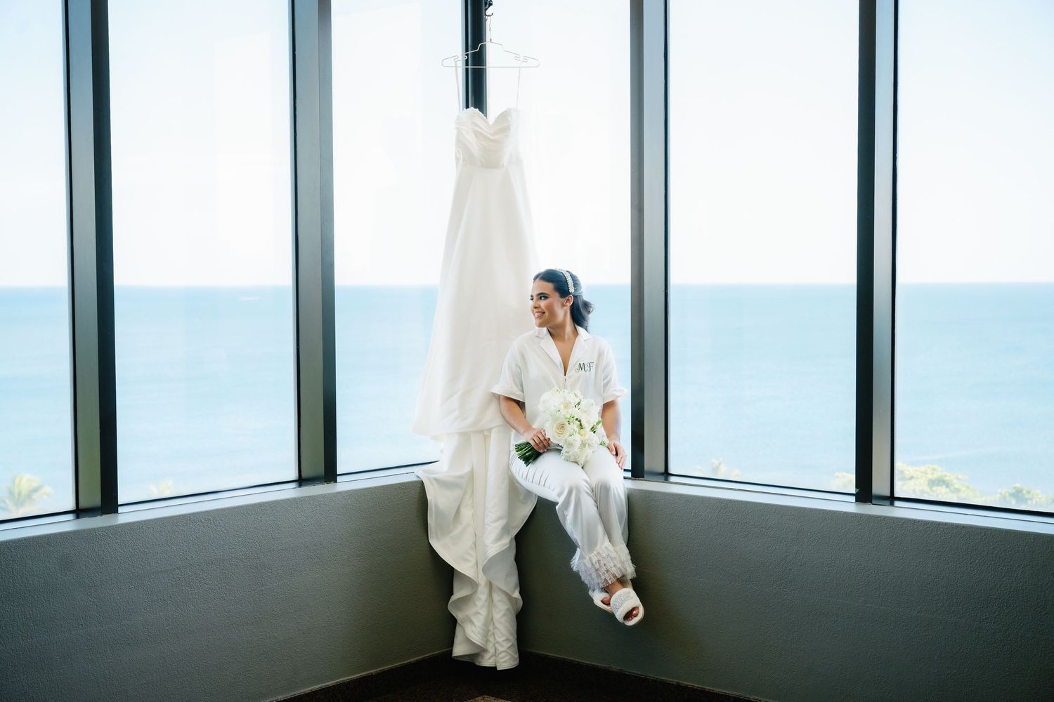 A bride in a white wedding dress stands by large windows overlooking a scenic ocean view.