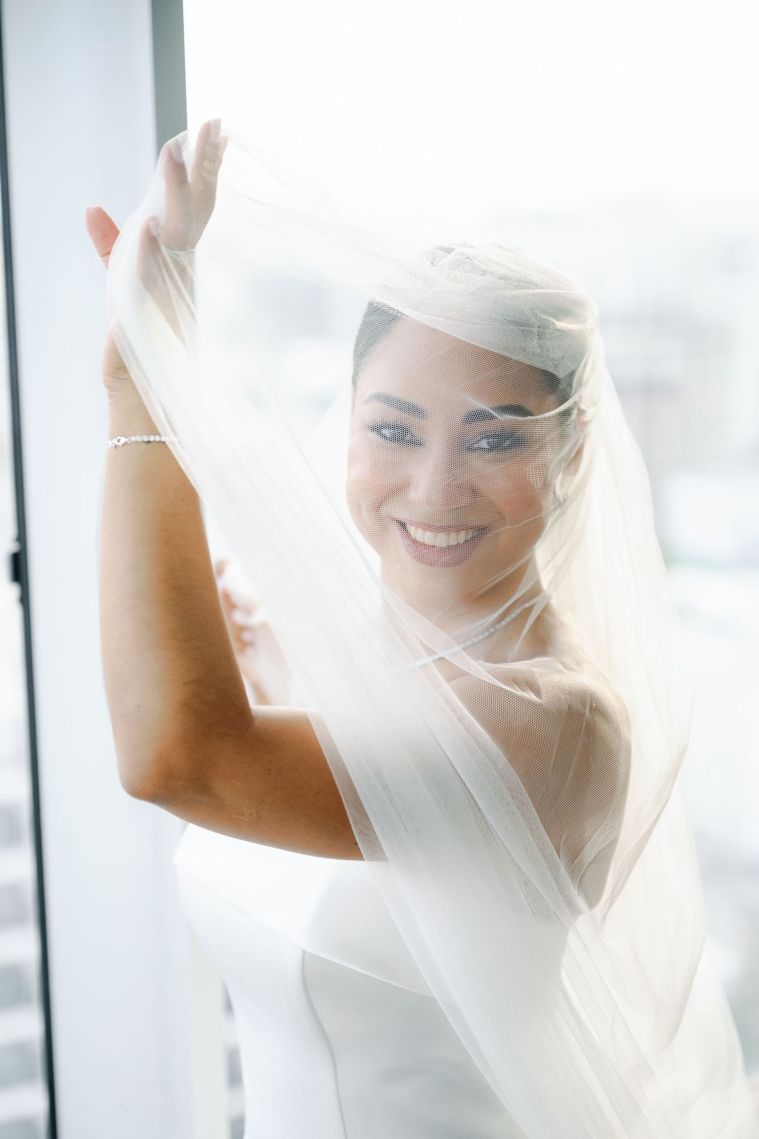 A bride in a white wedding dress and veil smiles joyfully in bright natural light near windows.
