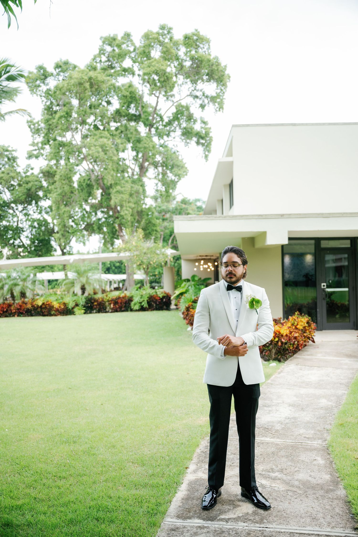 Ring bearer in white tuxedo jacket and black pants stands on outdoor walkway surrounded by green lawn.