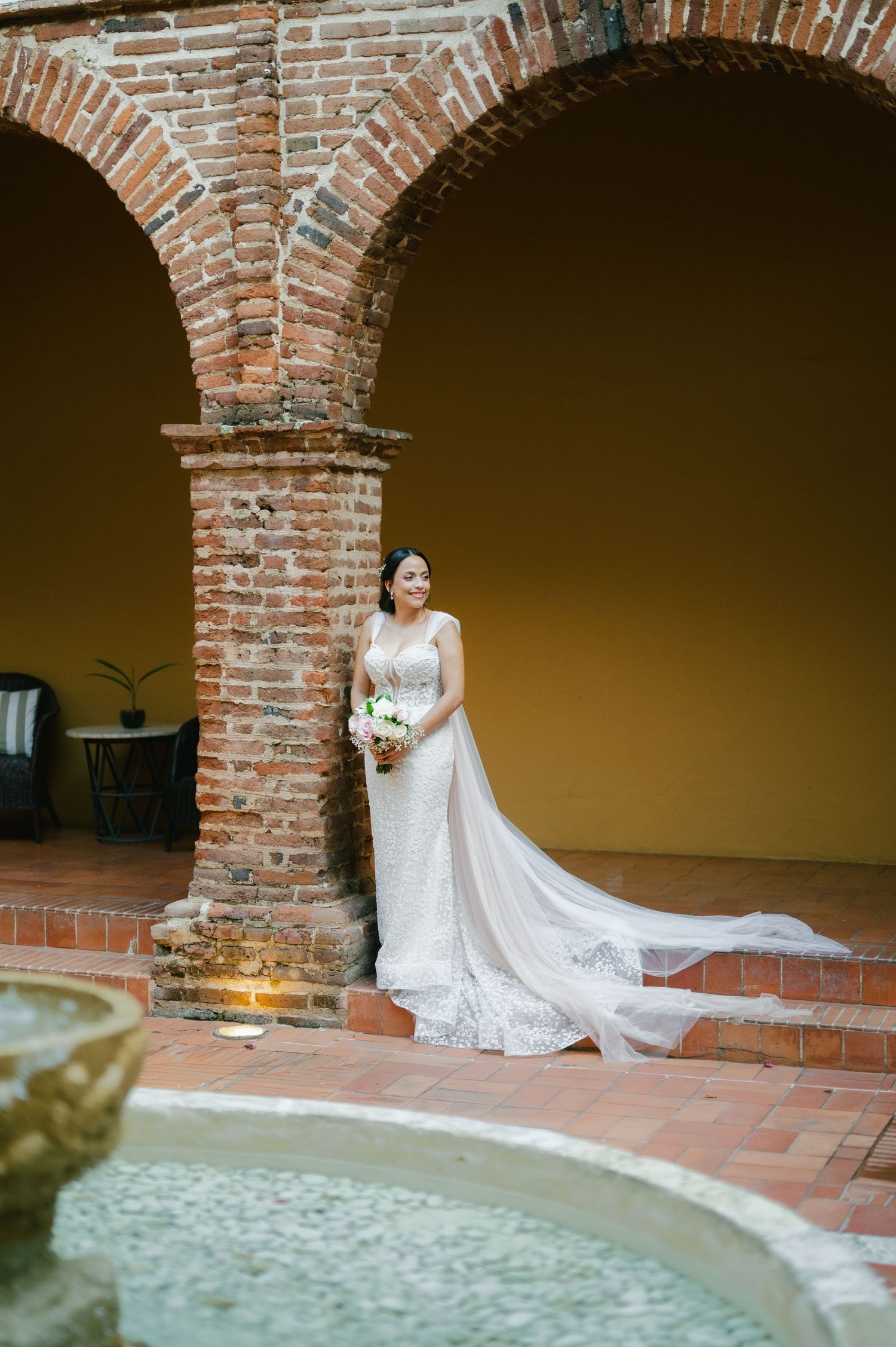 A bride in a flowing white lace wedding dress poses under rustic brick archways at a historic venue.