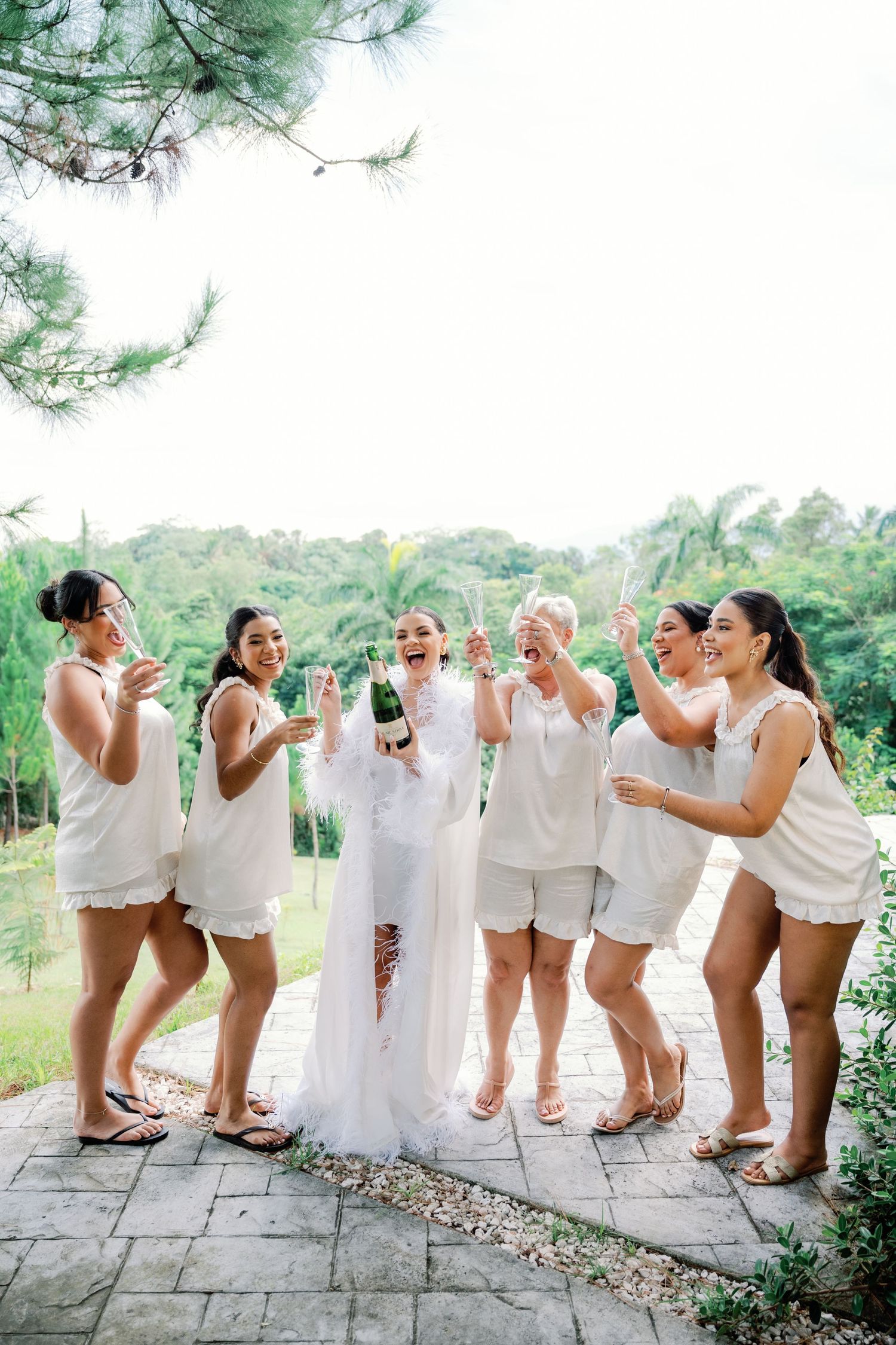 Wedding party in white outfits poses playfully on stone patio with scenic mountain backdrop.