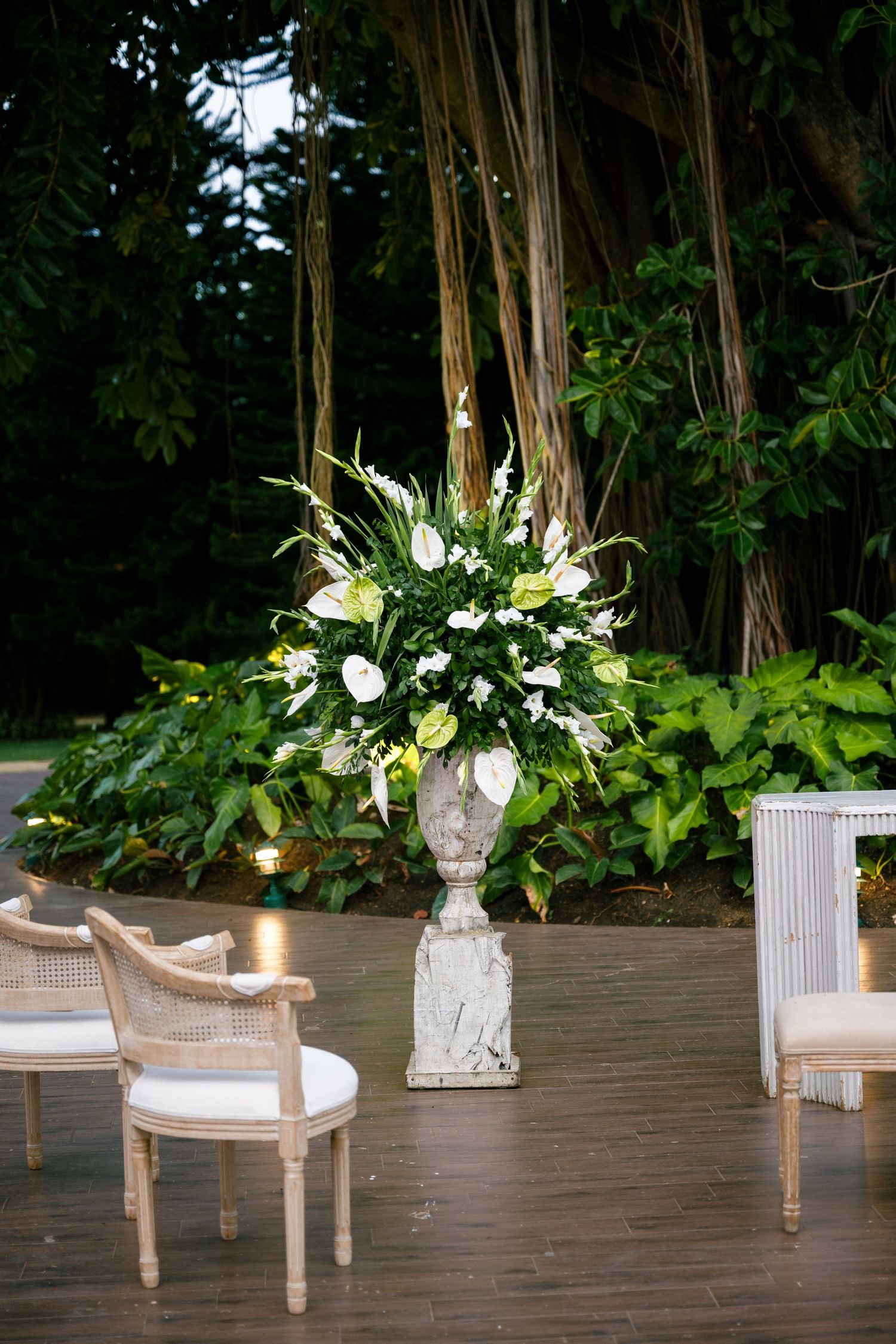 Elegant white floral arrangements in stone vases adorn a wooden deck with wicker chairs and ambient lighting at dusk.