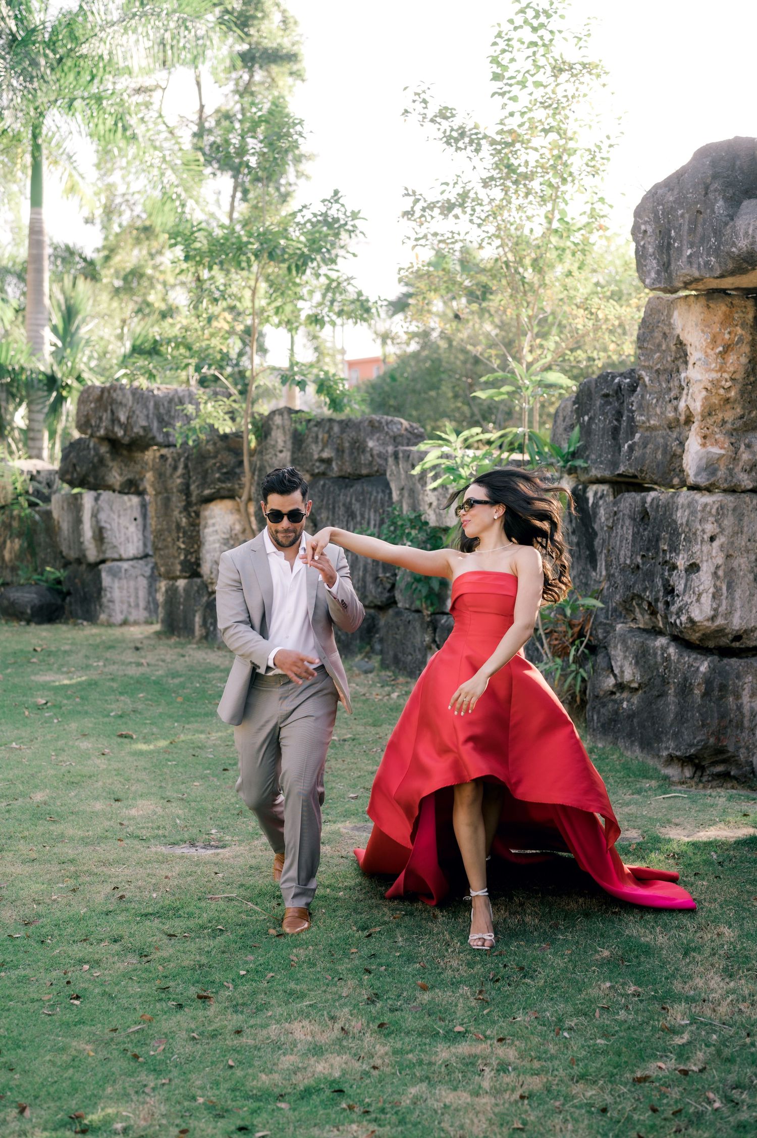 A couple in formal attire poses playfully against stone ruins in a garden setting with lush greenery.