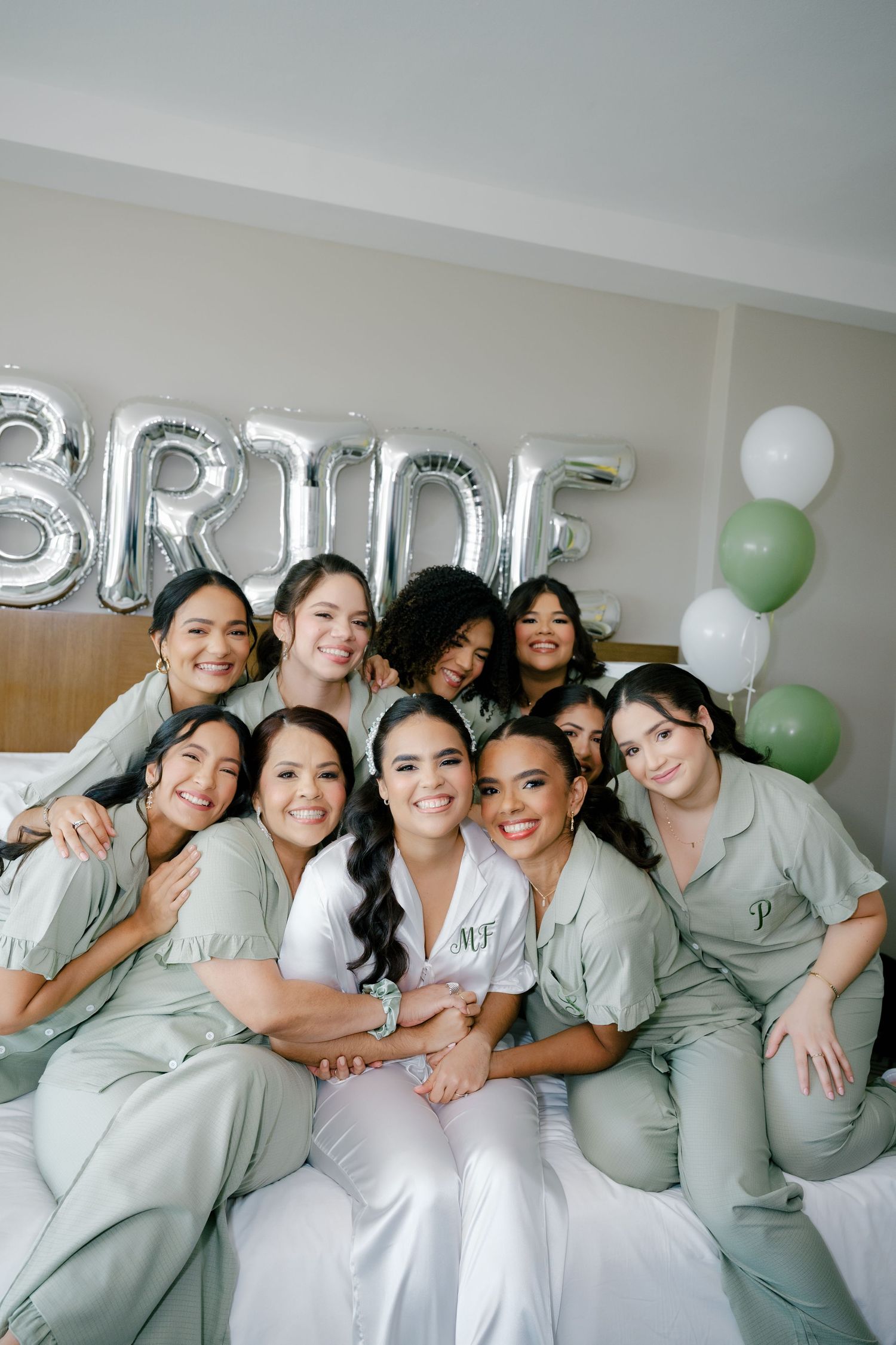 Wedding party wearing matching sage green outfits poses together on a couch with silver letter balloons in background.