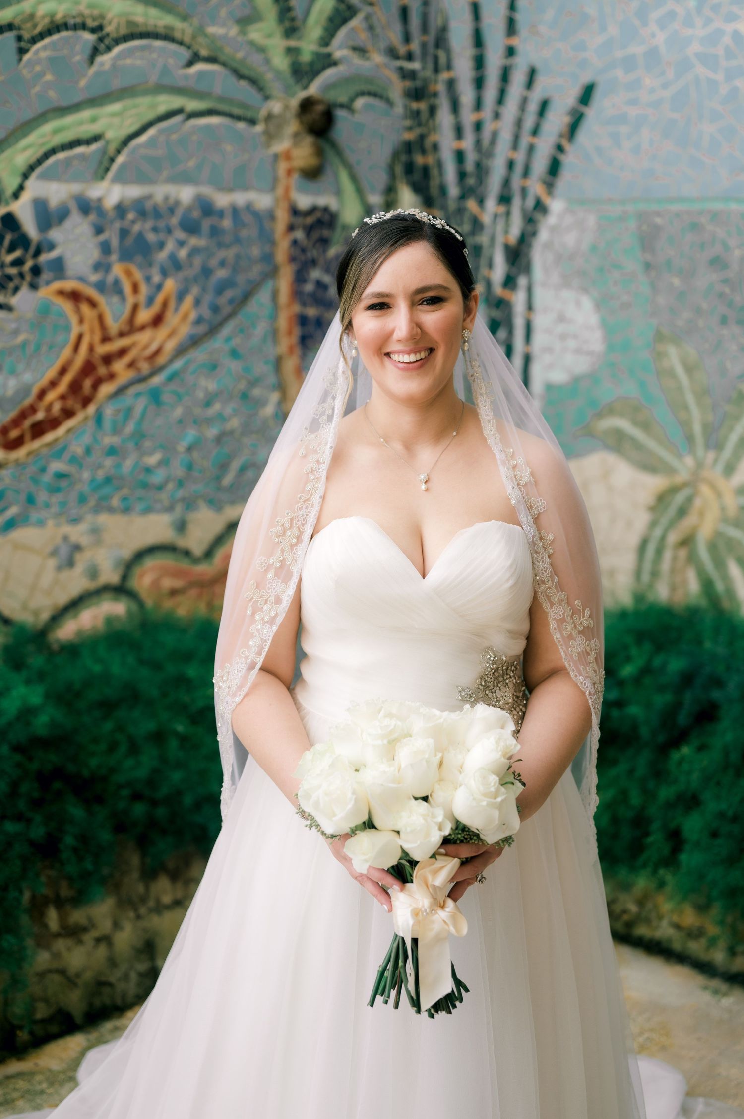 Elegant bride in white lace gown holds stunning white rose bouquet against a decorative garden backdrop.