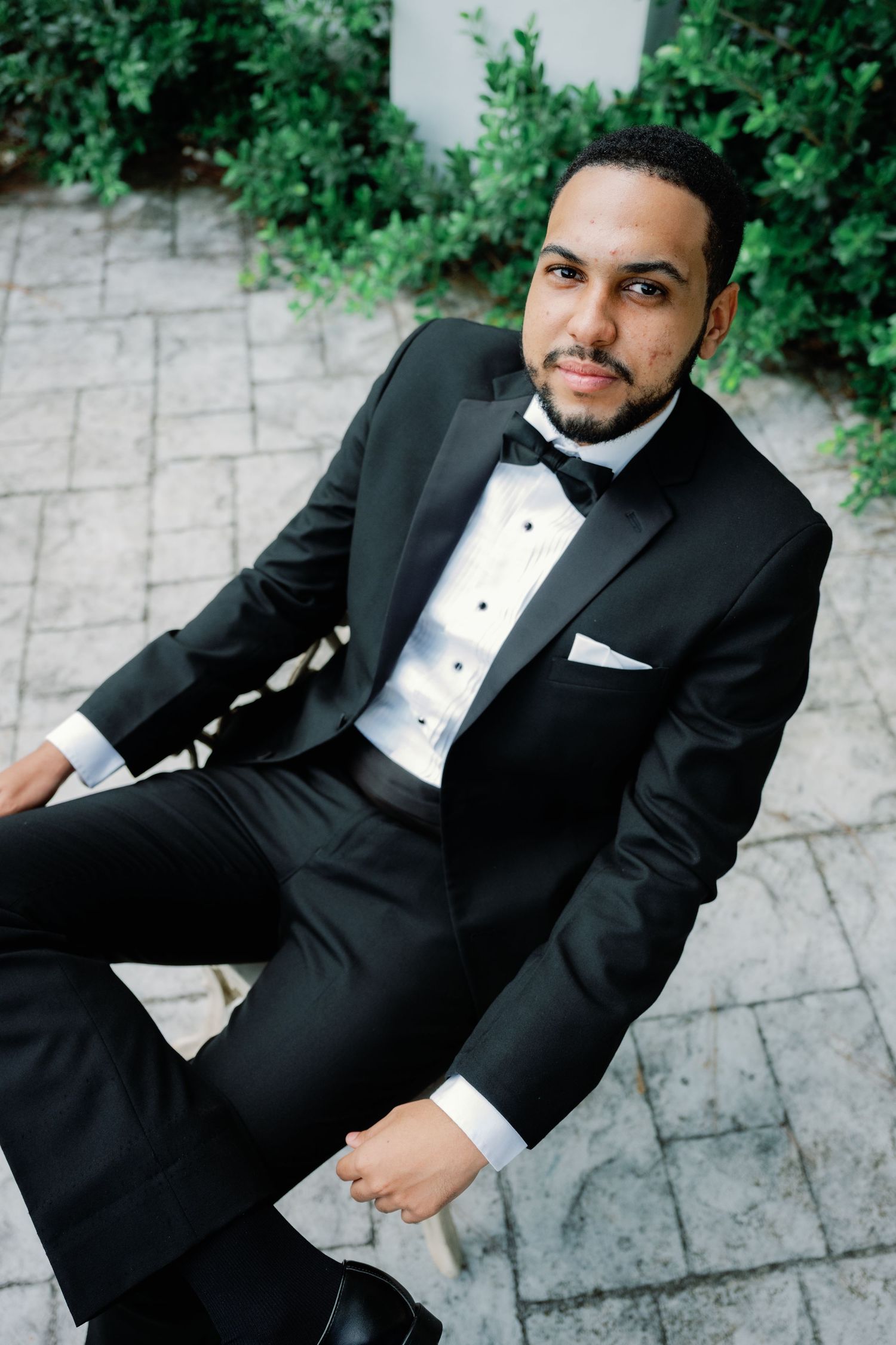 Groom in classic black tuxedo and bow tie poses outdoors against greenery.