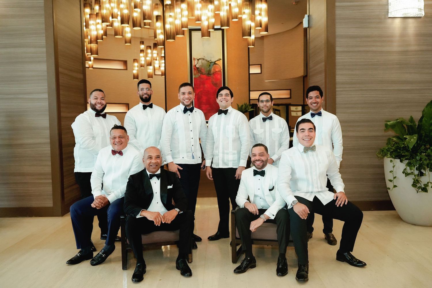 A group of groomsmen in white tuxedo jackets sit casually on a bench in an elegant hotel lobby.