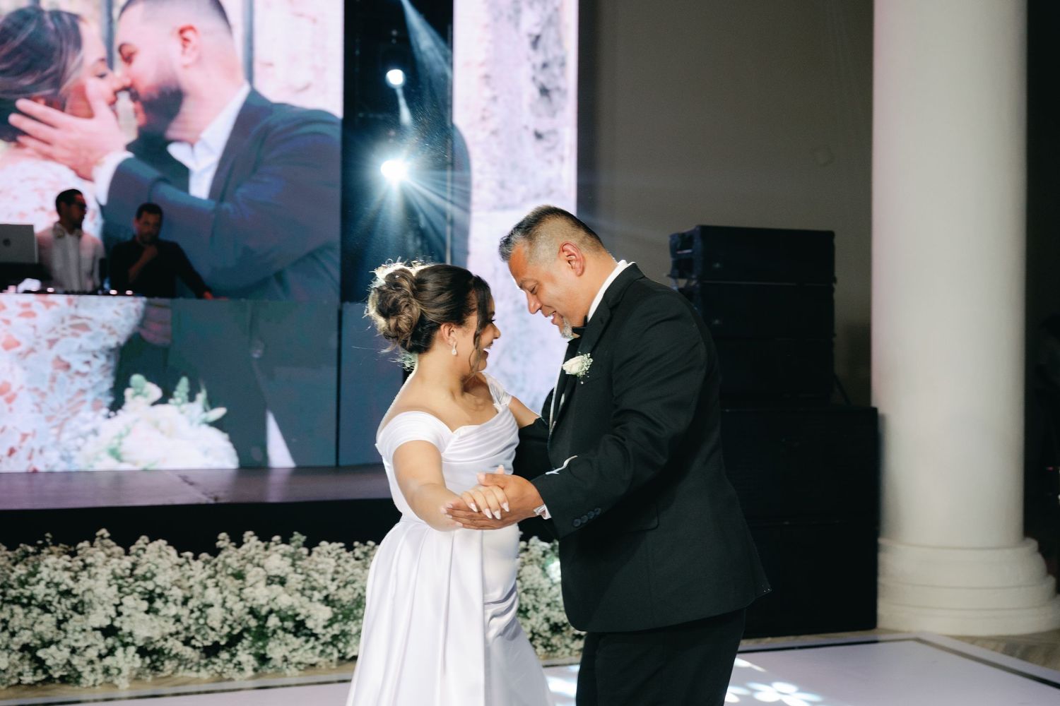 A couple shares their first dance at an elegant wedding reception surrounded by white floral decorations.