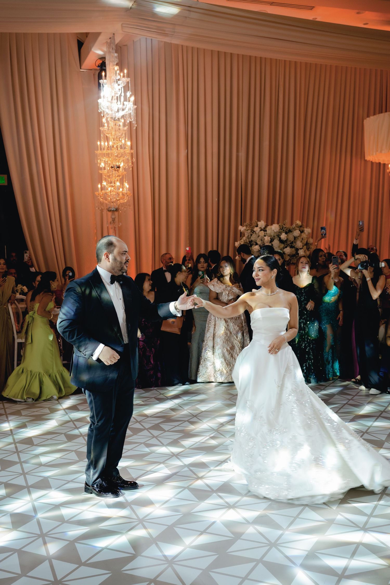 A couple shares their first dance on a geometric patterned dance floor under elegant crystal chandeliers at their wedding reception.