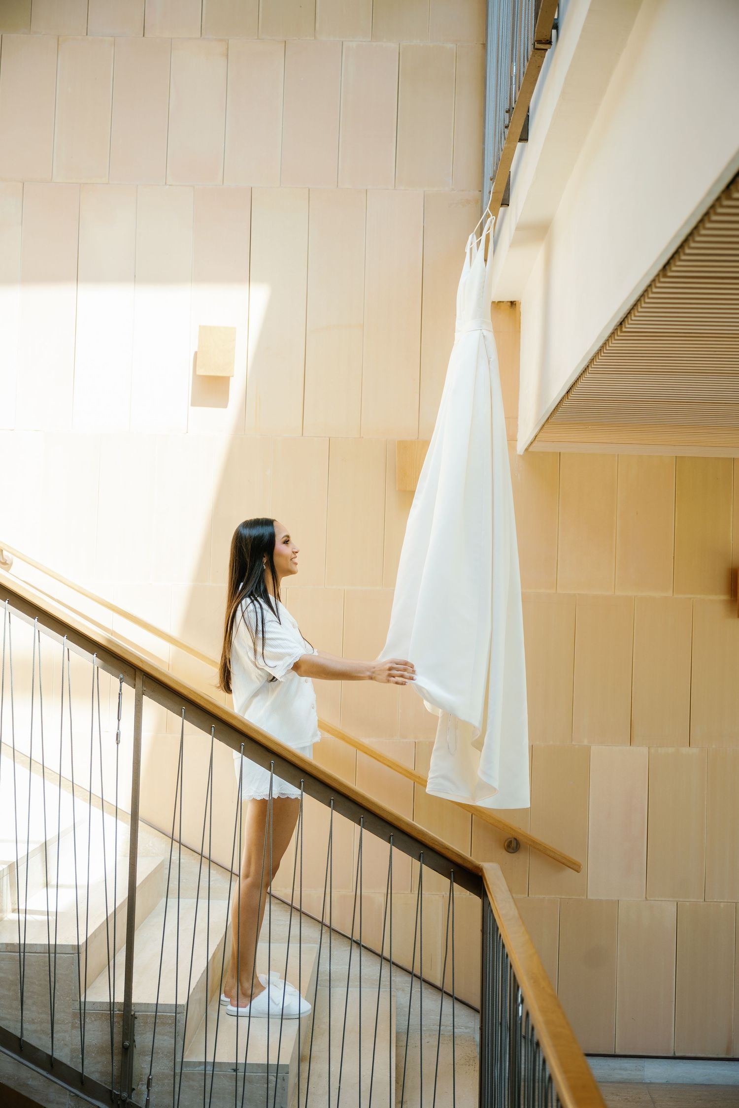 Person in white dress carries laundry up wooden staircase in bright sunlit interior.