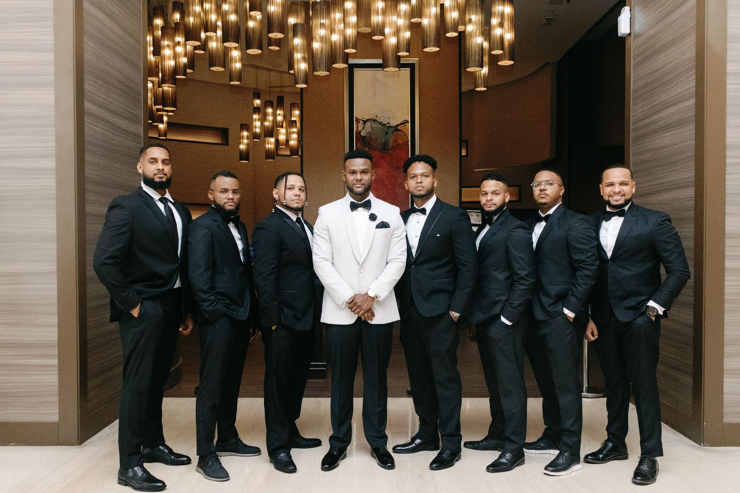 A group of groomsmen in black tuxedos sit together on steps under elegant chandeliers in a formal venue.