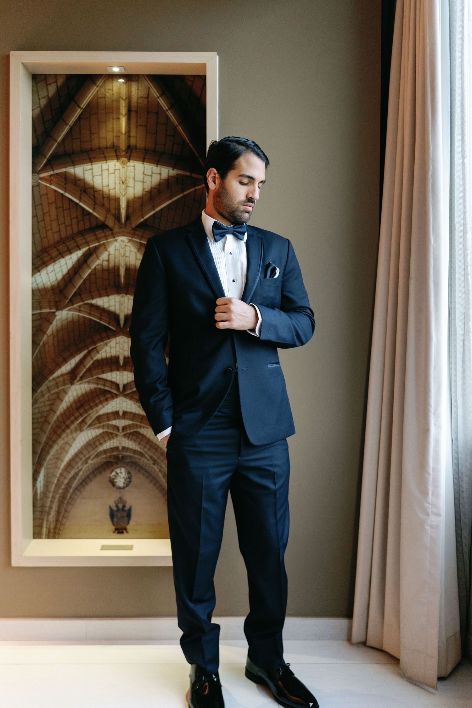 A groom in a classic black tuxedo and bow tie stands by a window in an elegant hotel room.