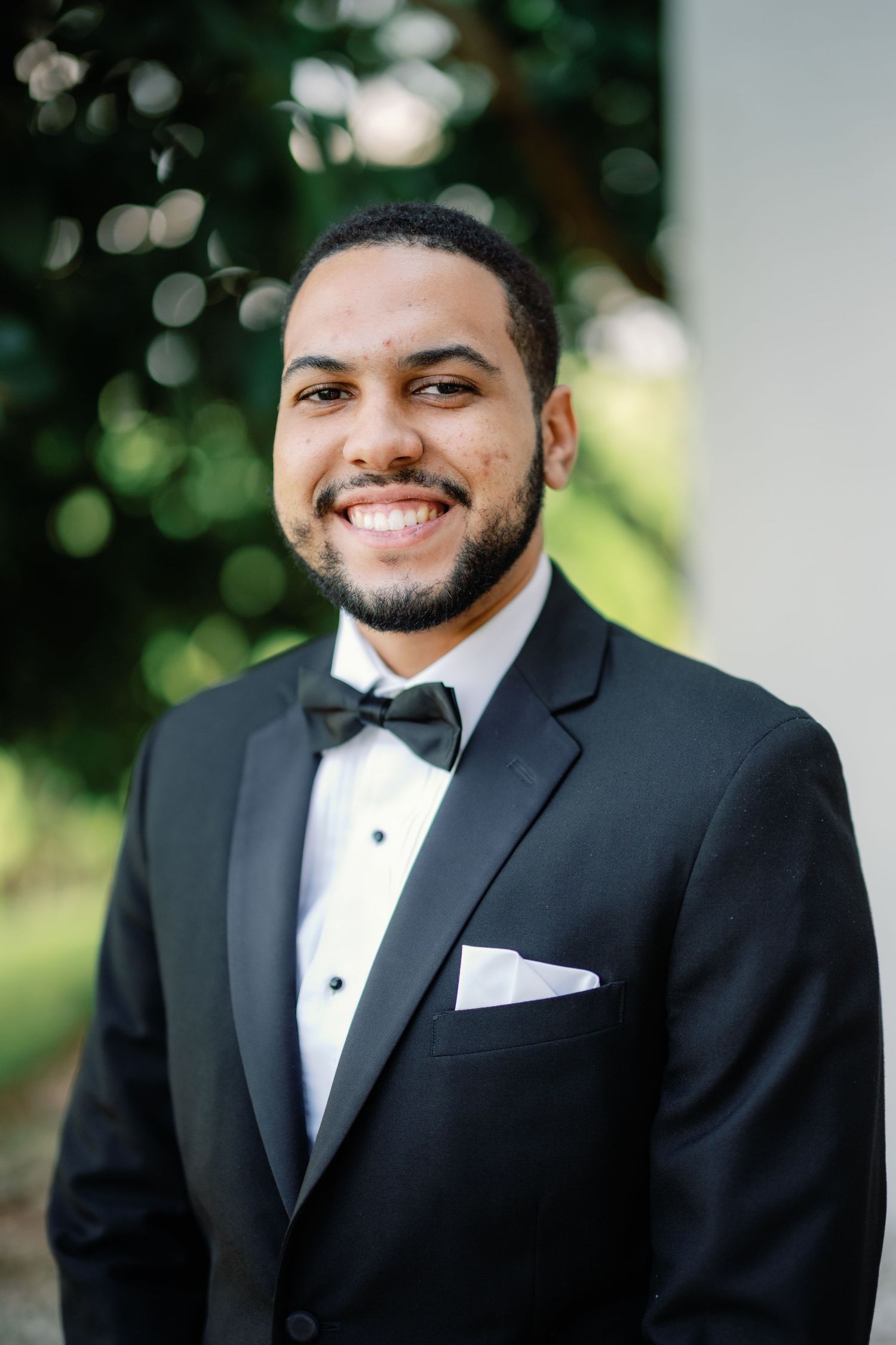 A portrait of a groom in a classic black tuxedo and bowtie smiling outdoors.