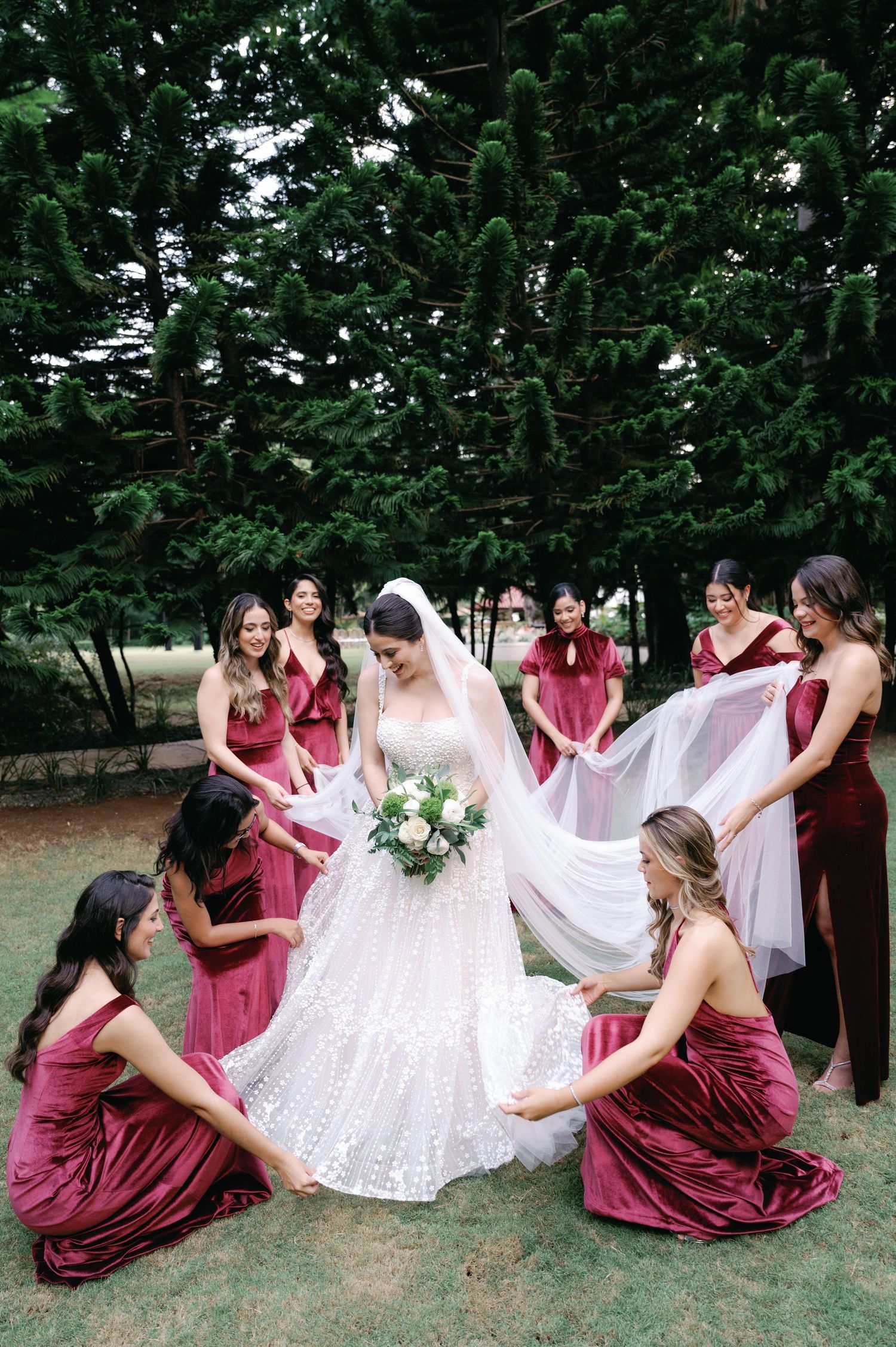 Bridesmaids in burgundy dresses help arrange the bride's wedding gown outdoors.