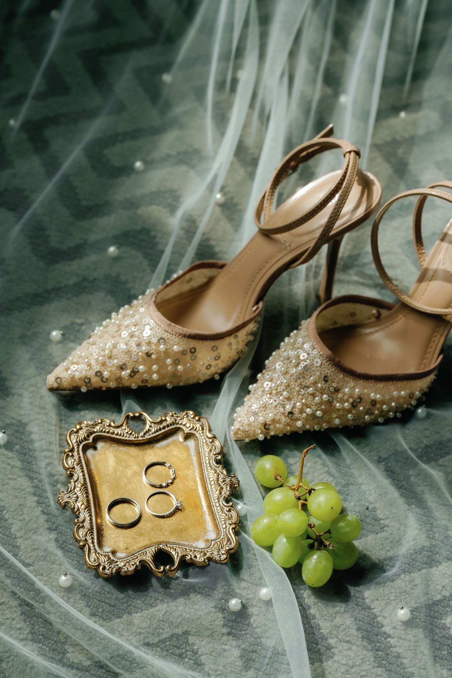 Elegant pearl-studded bridal heels next to vintage gold frame with wedding rings and fresh green grapes.