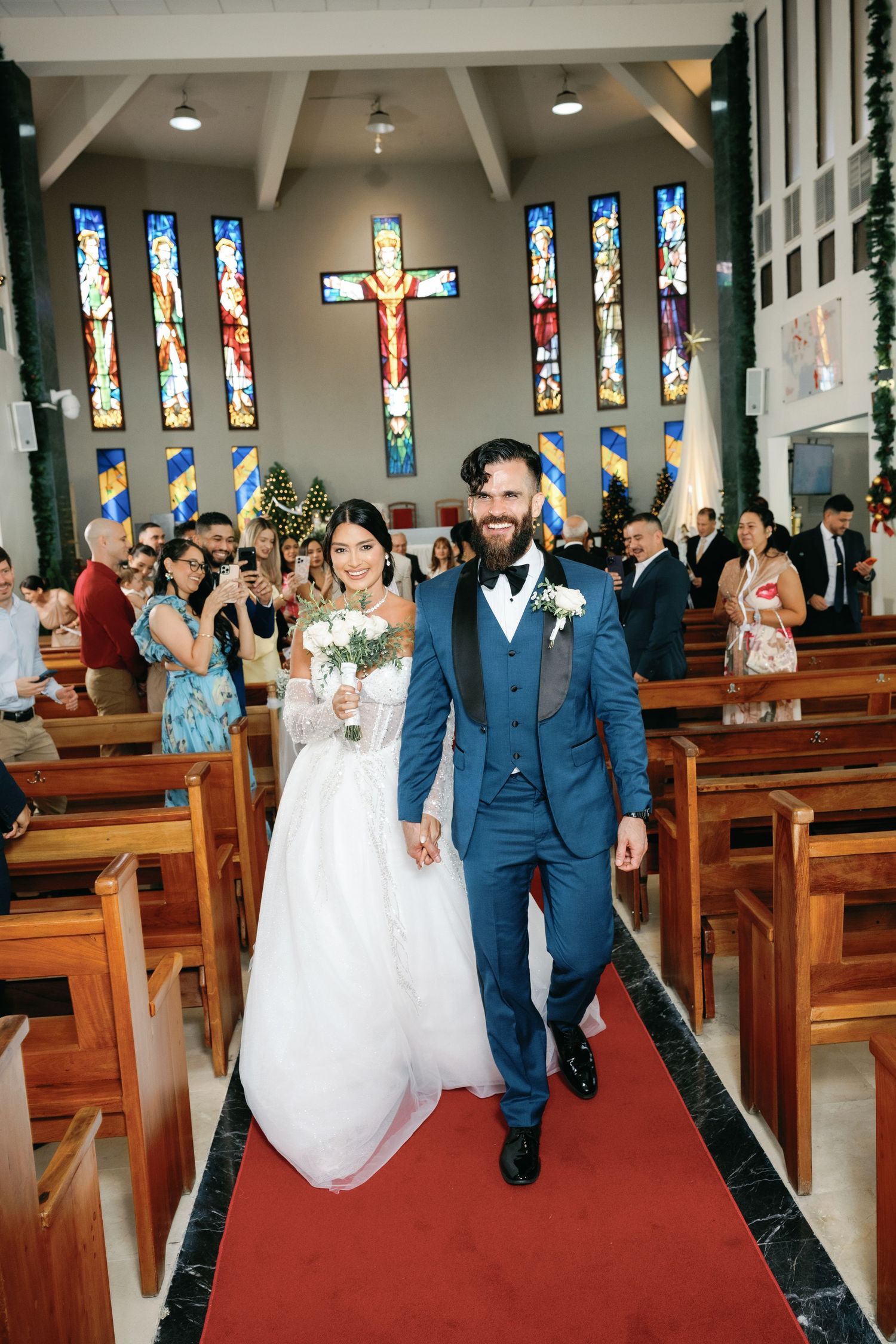 A couple walks down the church aisle after their wedding ceremony with stained glass windows and crucifix in background.