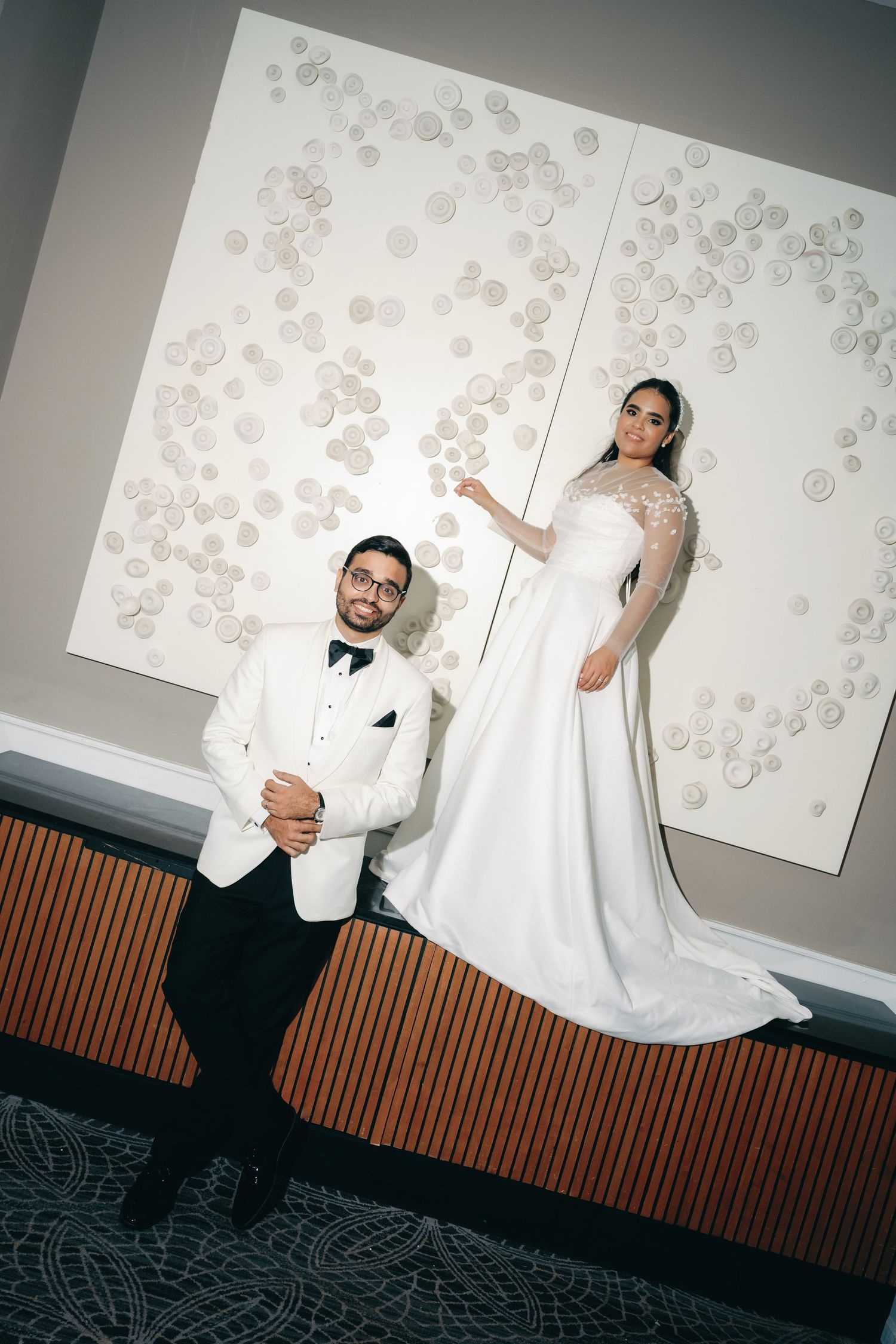 A couple poses in elegant white wedding attire against a wall decorated with delicate floral artwork.