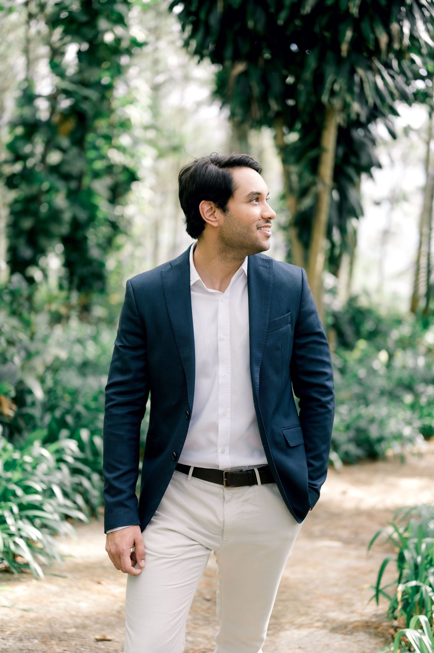 A person wearing a navy blazer and white pants walks along a tree-lined path in a natural outdoor setting.